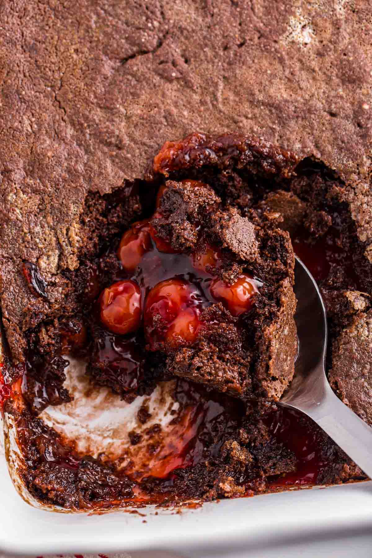 Spoon lifting a portion of cherry dump cake from a baking dish showing chocolate cake and cherry filling inside.