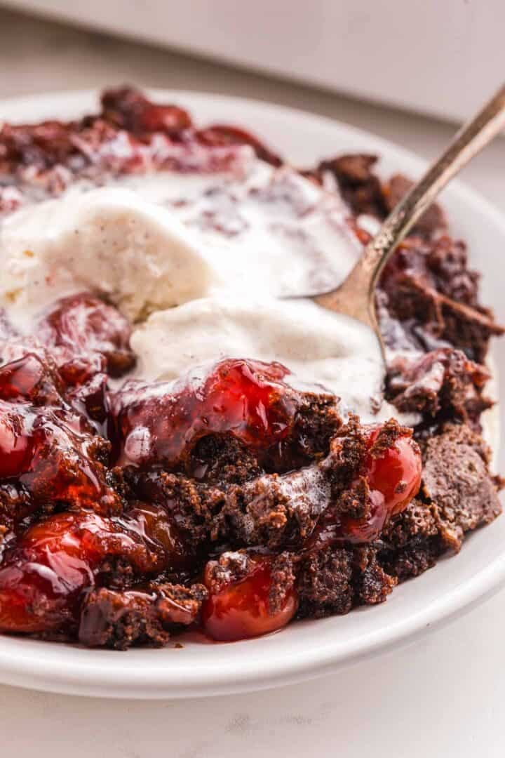 Close up of cherry dump cake with vanilla ice cream and a spoon cutting into the chocolate cake and cherries.