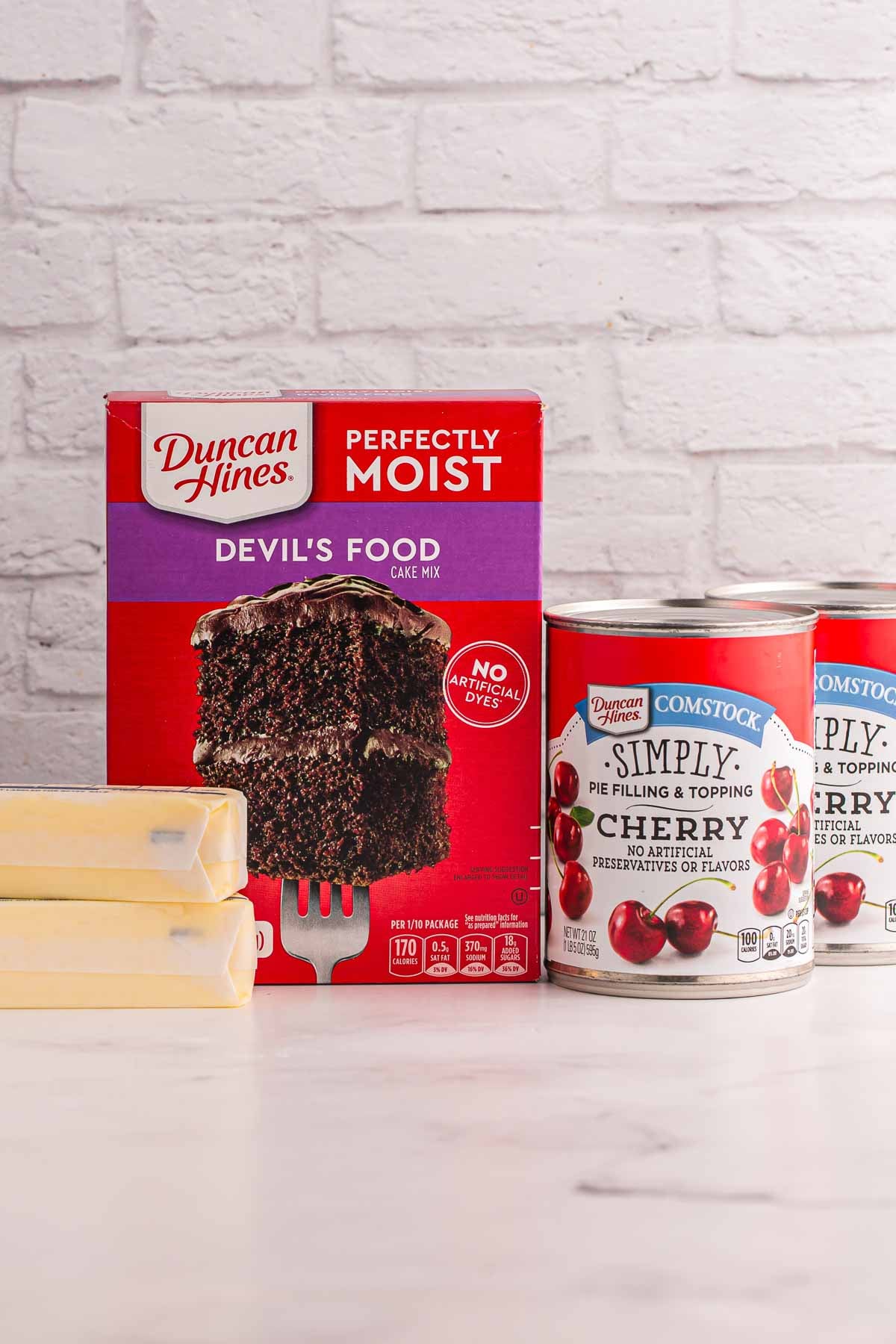 Ingredients for cherry dump cake including Duncan Hines Perfectly Moist Devil's Food Cake Mix and two cans of cherry pie filling with two sticks of butter.