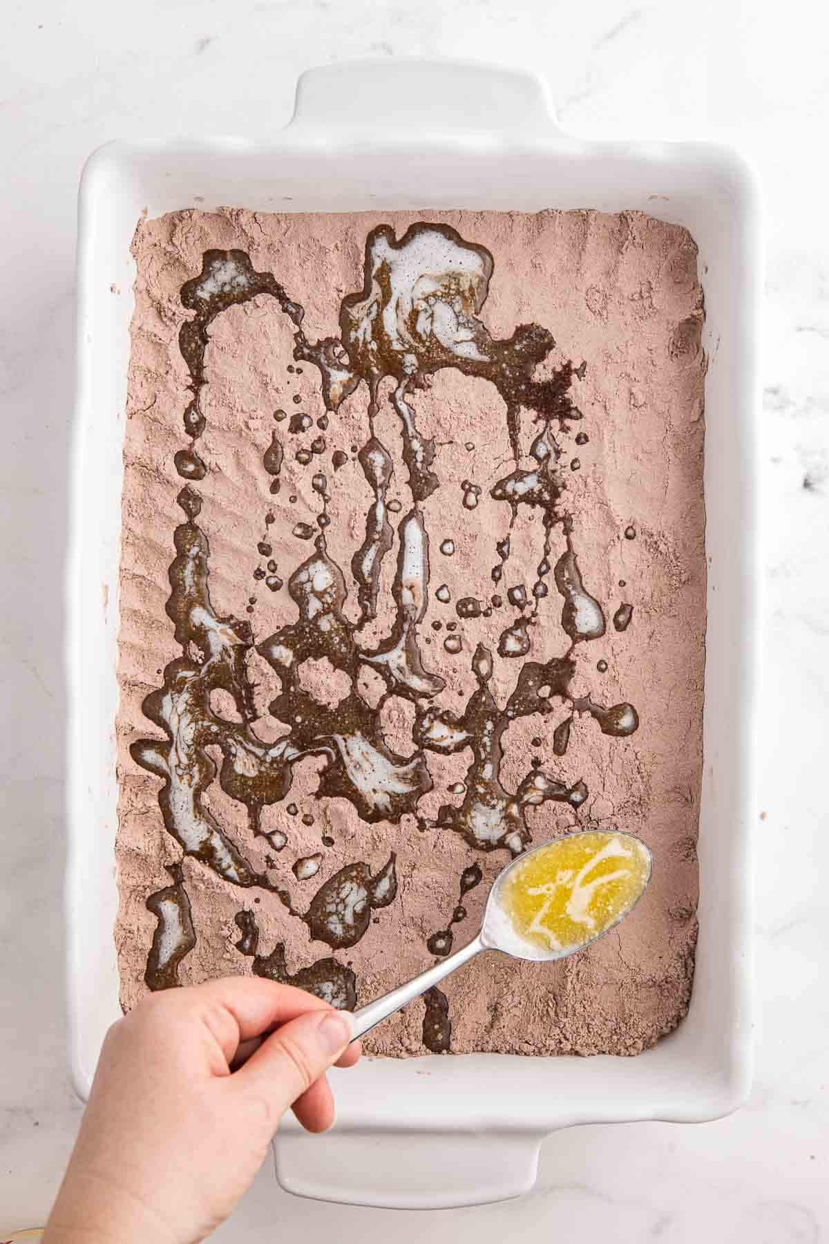Melted butter being spooned over dry cake mix in a baking dish by a hand holding a spoon.