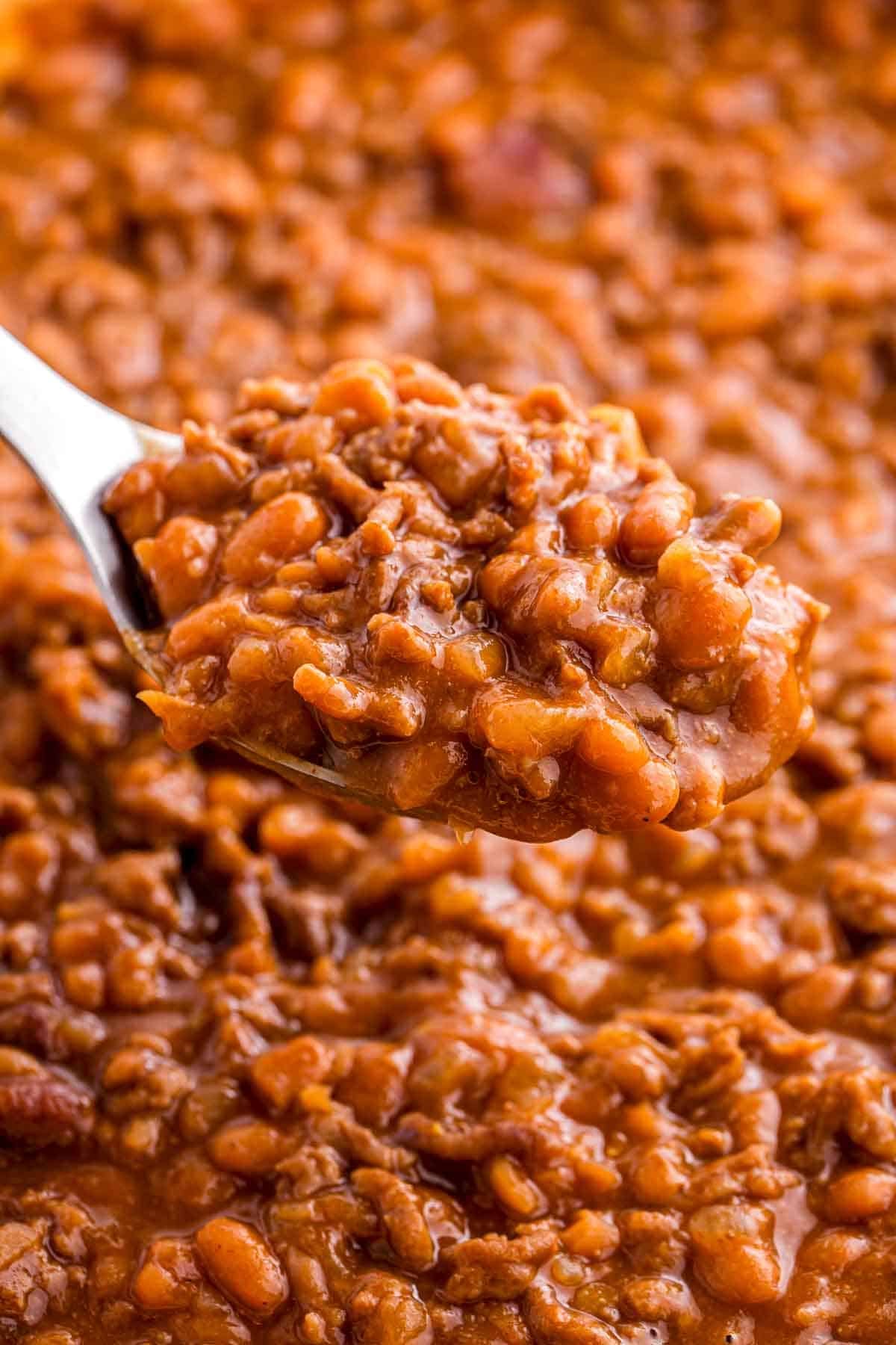 Spoon lifting a thick scoop of baked beans with ground beef from a pot. The close up highlights the saucy texture, tender beans, and crumbled beef.