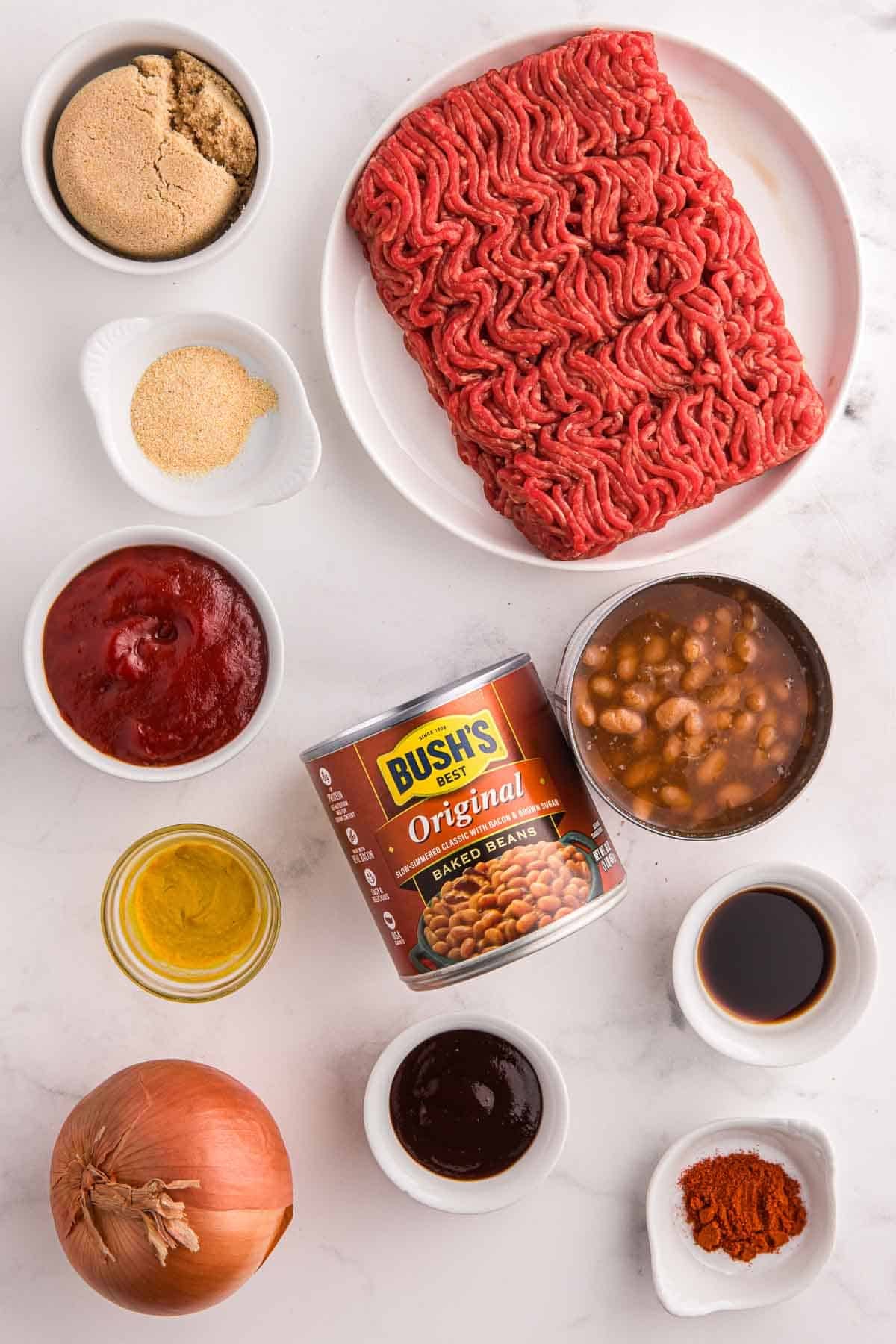 Ingredients arranged for baked beans with ground beef including raw ground beef, diced yellow onion, canned baked beans, brown sugar, ketchup, BBQ sauce, yellow mustard, Worcestershire sauce, smoked paprika, and garlic powder on a light surface.