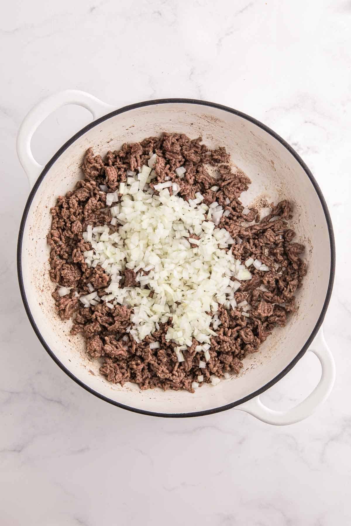 Ground beef browning in a large white pot topped with finely diced onion. The image shows the first cooking step for making baked beans with ground beef.