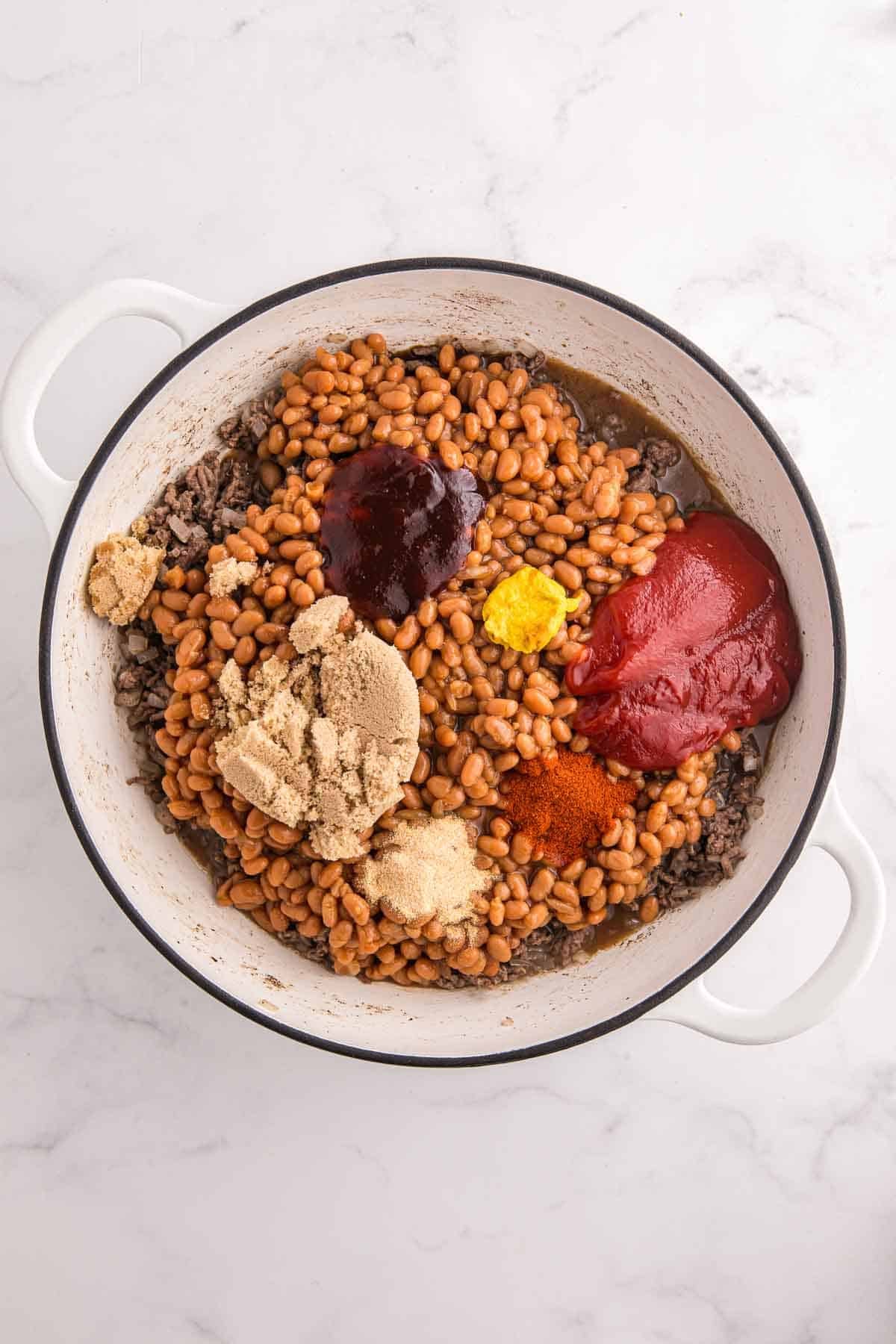 Large pot filled with cooked ground beef, baked beans, brown sugar, ketchup, BBQ sauce, mustard, smoked paprika, garlic powder, and seasonings before stirring. The ingredients are layered together to make baked beans with ground beef.