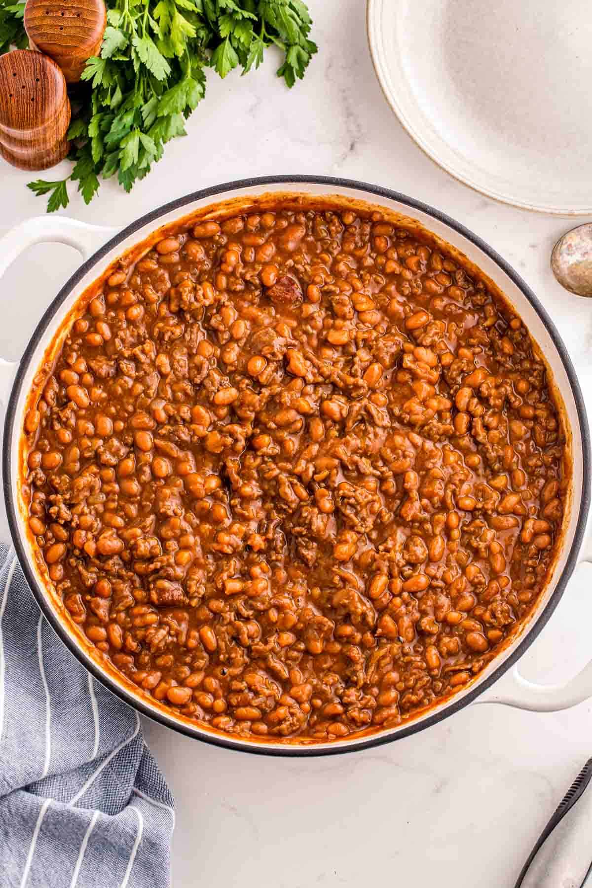 Overhead view of a large pan filled with baked beans and ground beef in a thick savory sauce. Fresh parsley and serving utensils surround the dish on a light countertop.