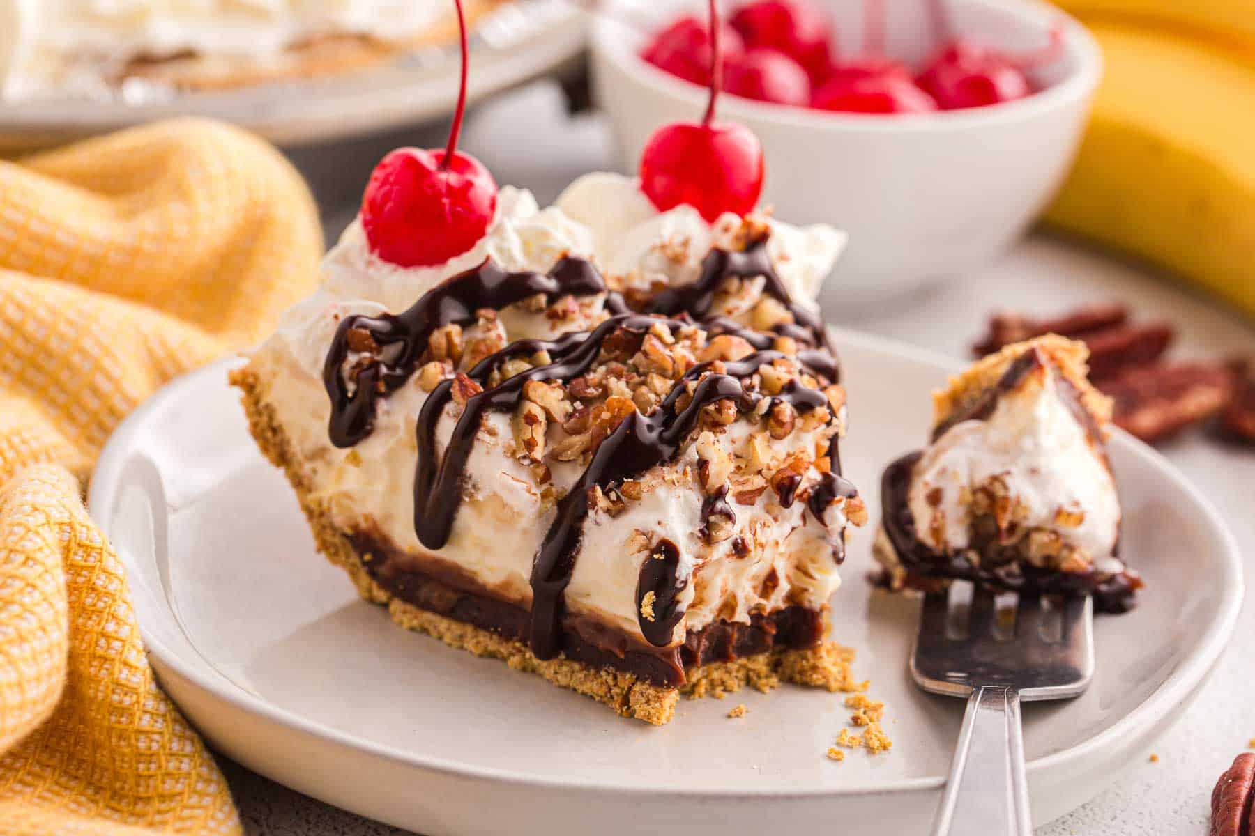 Slice of no bake banana split pie topped with whipped cream, chopped pecans, chocolate syrup, and cherries served on a plate with a forkful beside it.