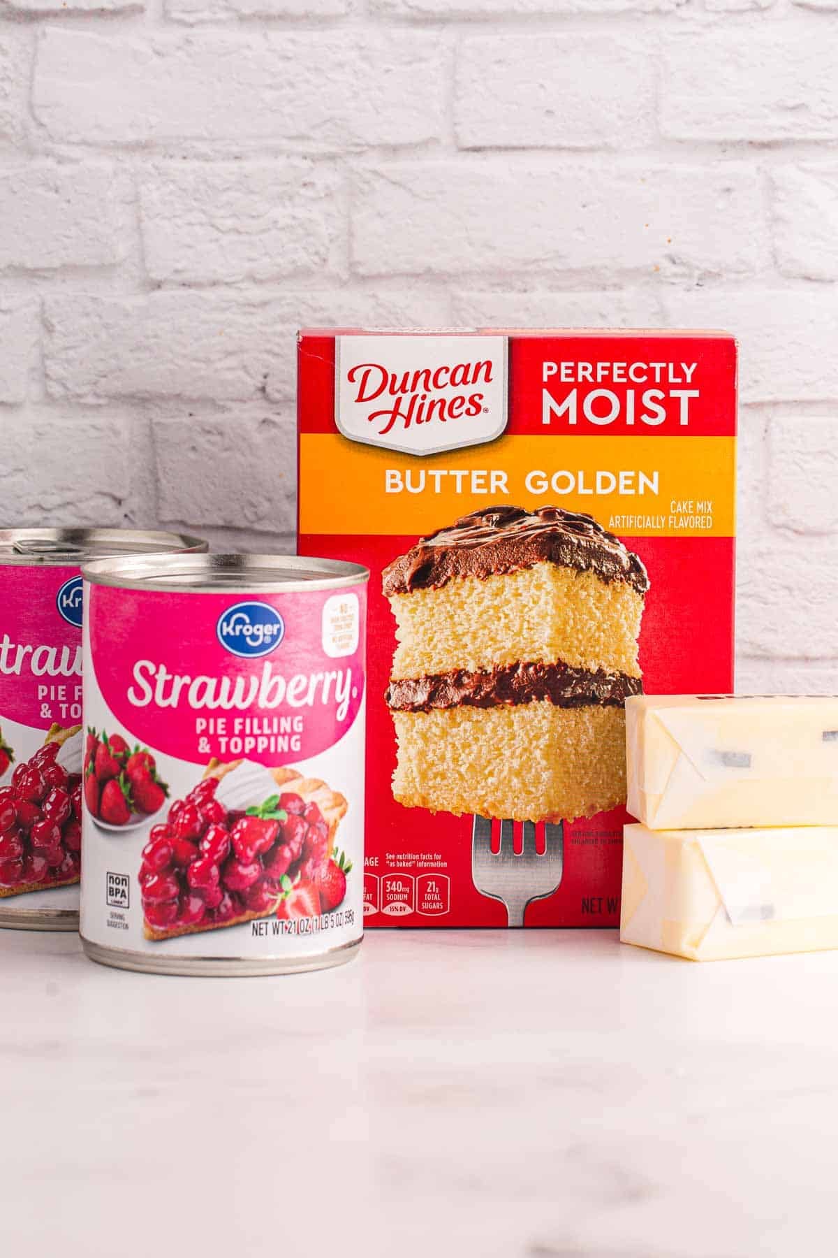Ingredients for strawberry dump cake including strawberry pie filling, yellow cake mix, and butter.