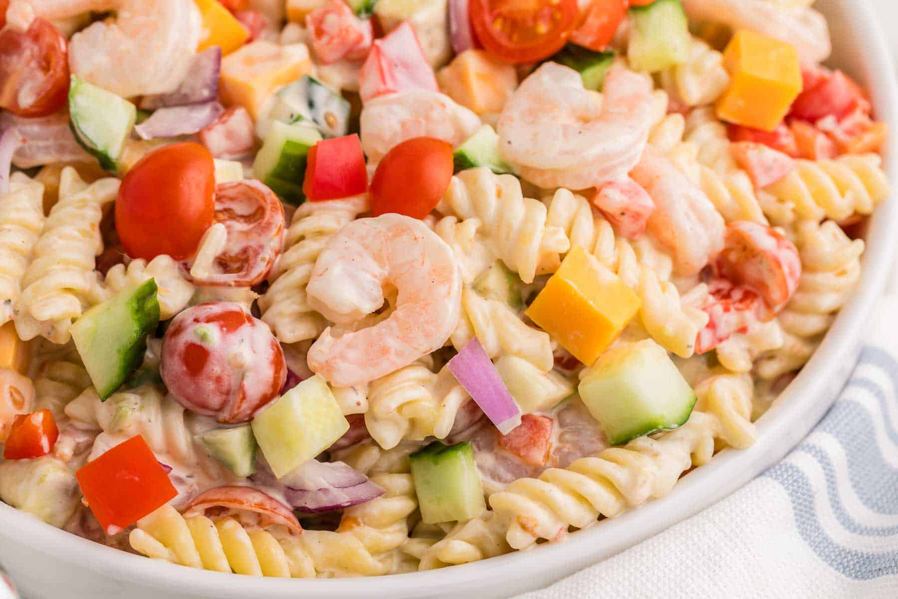 Close up of shrimp pasta salad with rotini pasta, shrimp, diced cucumber, cherry tomatoes, red bell pepper, red onion, and cheddar cheese mixed in a creamy dressing.