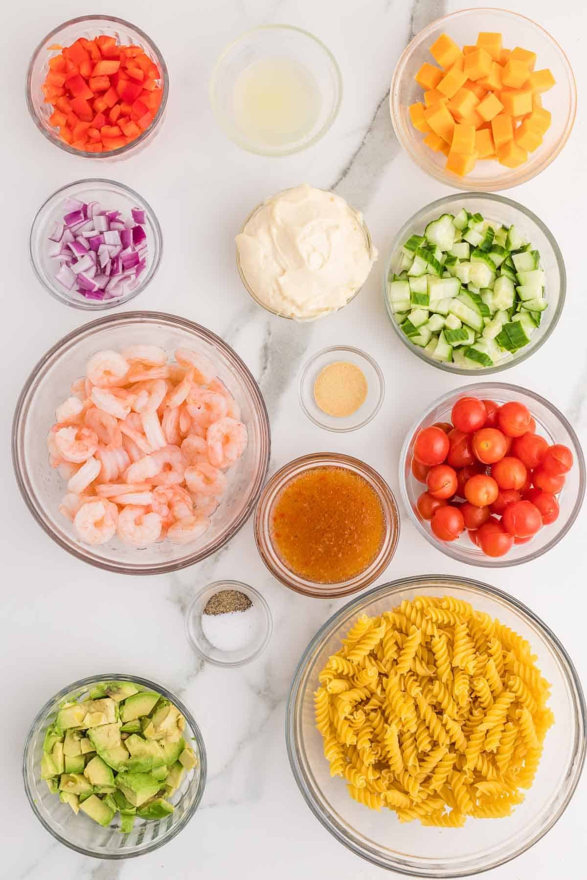 Ingredients for shrimp pasta salad arranged separately, including cooked shrimp, rotini pasta, cherry tomatoes, cucumber, avocado, red bell pepper, red onion, cheddar cheese, mayonnaise, Italian dressing, lemon juice, garlic powder, salt, and black pepper.