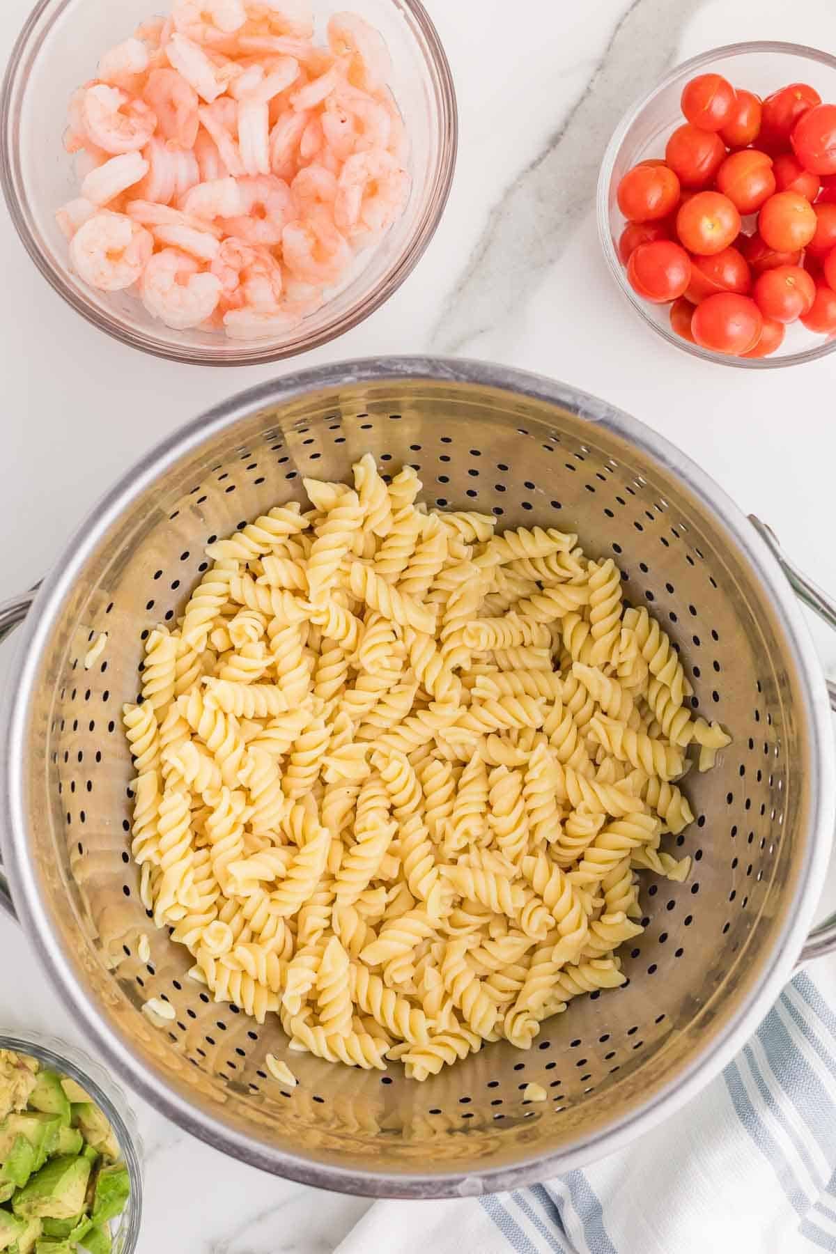 Cooked rotini pasta drained in a colander with bowls of shrimp and cherry tomatoes prepared for shrimp pasta salad.
