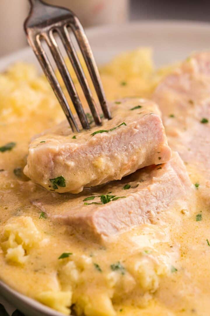 A fork lifts a piece of tender pork chop from a creamy ranch sauce. Mashed potatoes and another sauced pork chop fill the blurred background.