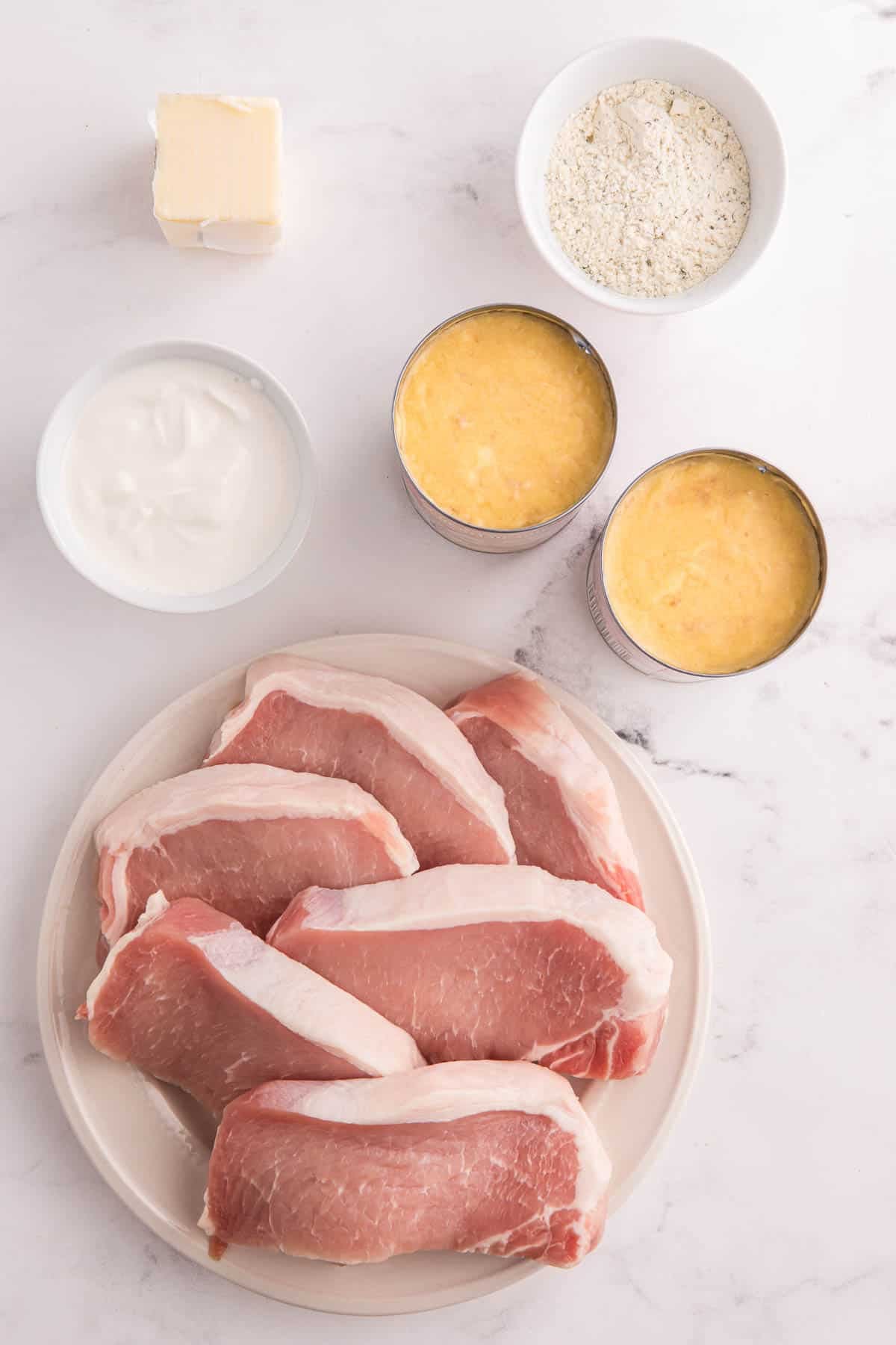 Ingredients for crockpot ranch pork chops arranged on a marble counter. Raw boneless pork chops sit beside cream of chicken soup. sour cream. ranch seasoning. and melted butter.
