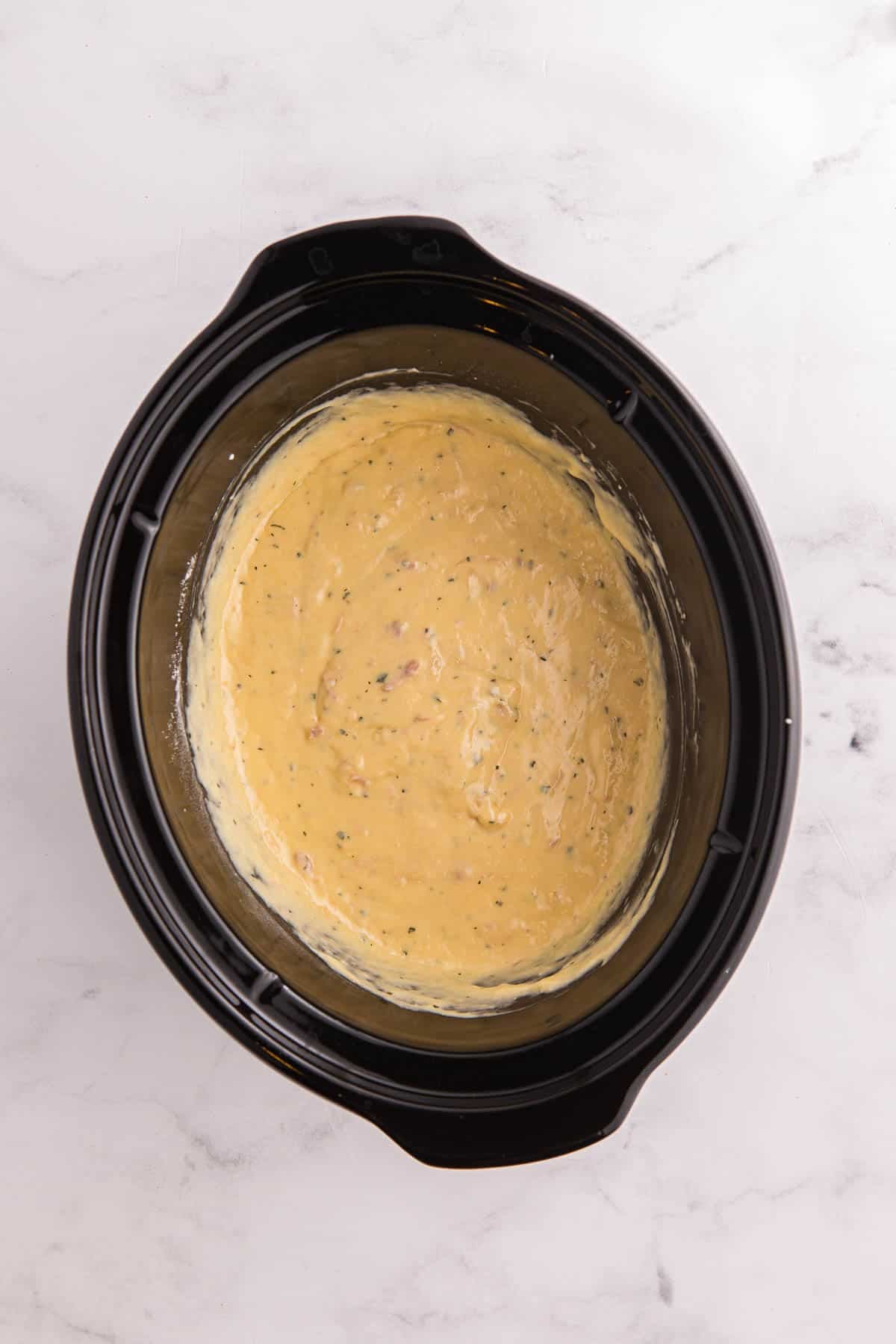 Cream of chicken soup. sour cream. ranch seasoning. and melted butter are mixed into a smooth sauce inside a black slow cooker. The sauce forms the creamy base for the pork chops.