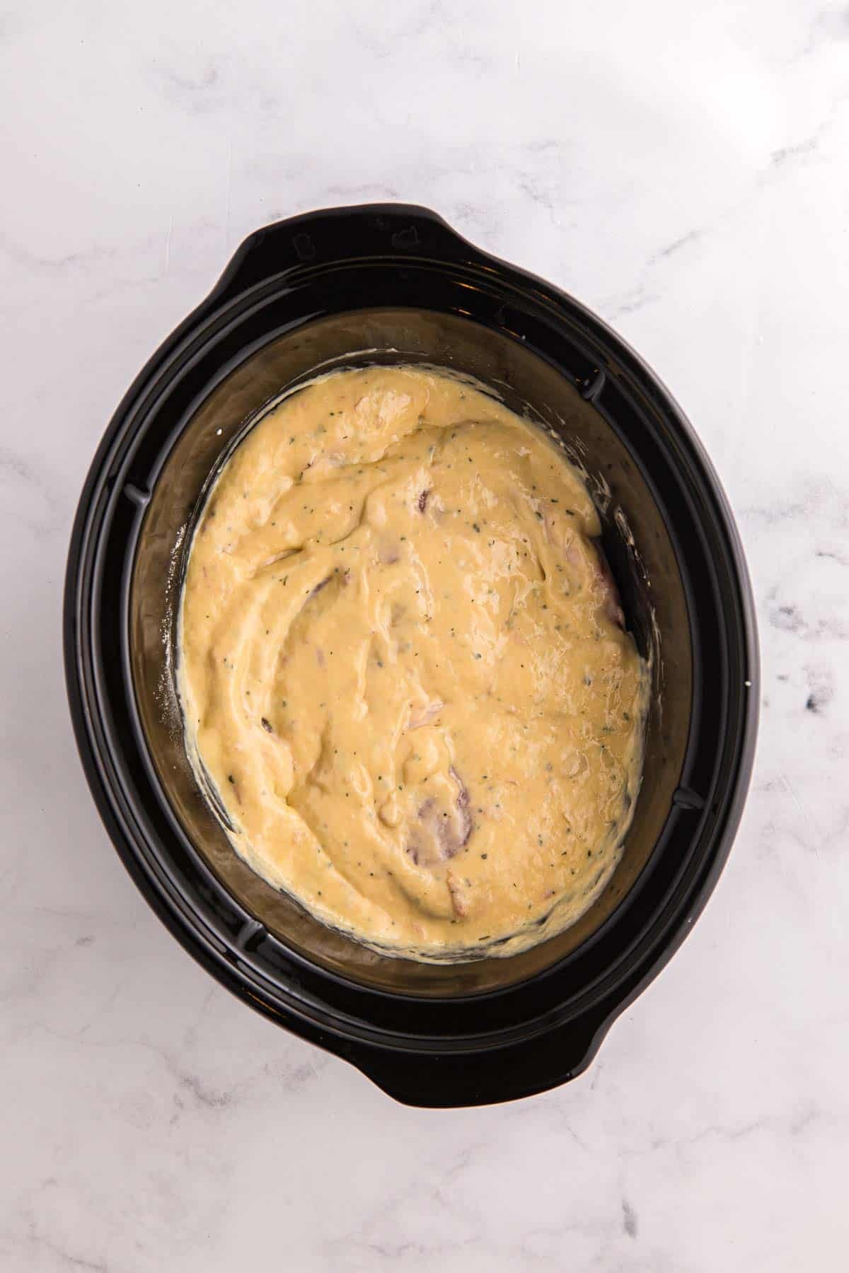 Raw pork chops are placed in a black slow cooker and coated with creamy ranch sauce. The chops overlap slightly under the thick sauce.