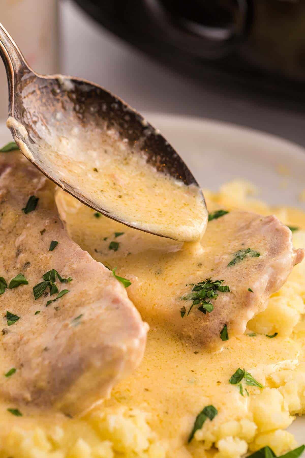 A spoon pours creamy ranch sauce over pork chops served on mashed potatoes. Fresh parsley adds color to the finished dish.