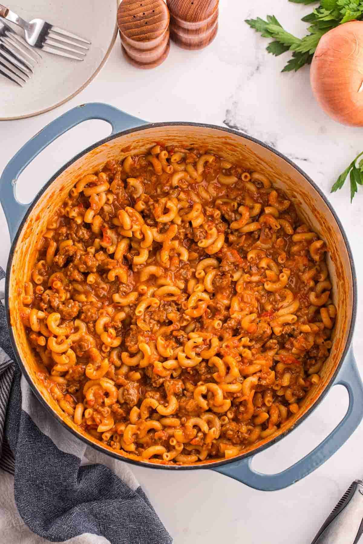 American goulash fills a blue handled Dutch oven on a white counter. The overhead view shows thick tomato sauce coating the ground beef and elbow macaroni.