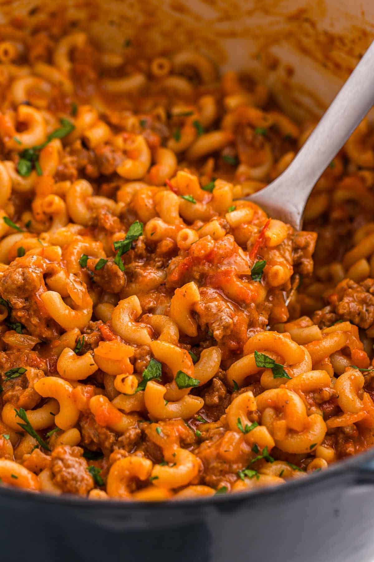 A serving spoon lifts saucy elbow macaroni and ground beef from the Dutch oven. Chopped parsley adds fresh green color to the rich tomato sauce.