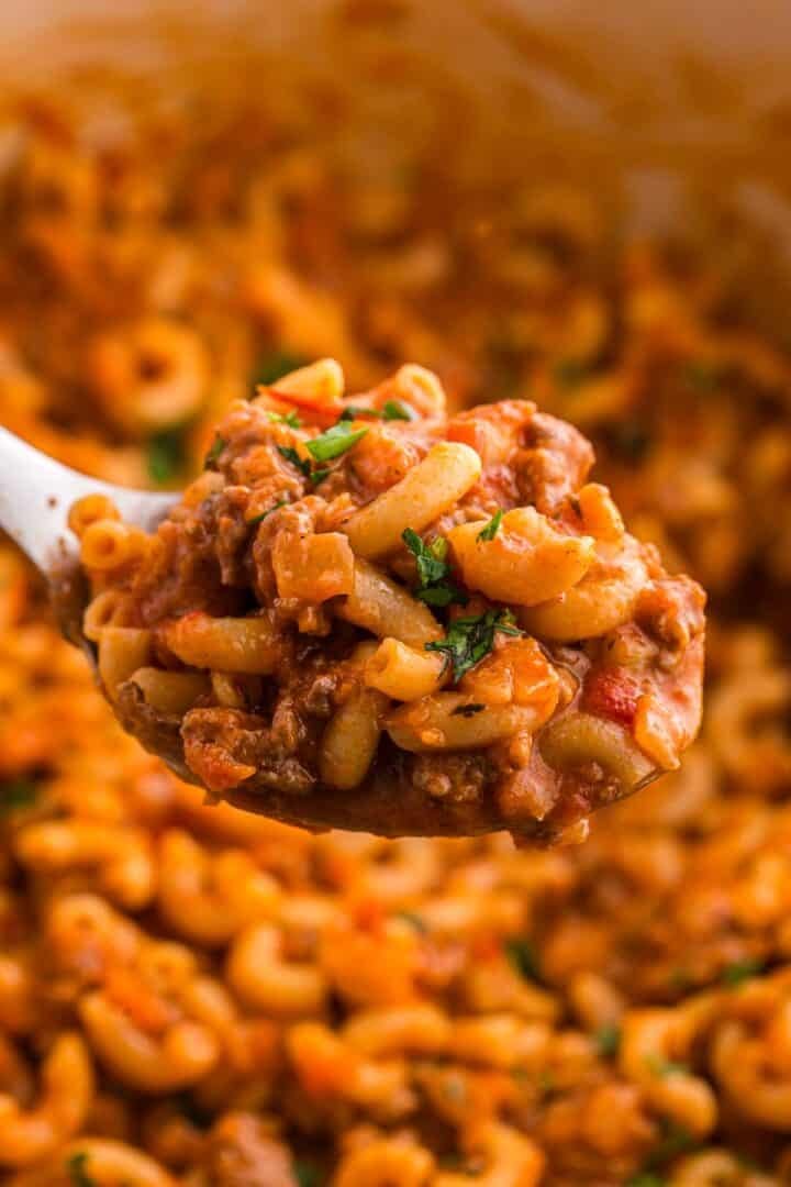 A heaping spoonful of ground beef elbow macaroni tomato sauce and chopped parsley is held above the pot. The close view highlights the thick and hearty texture of the finished dish.
