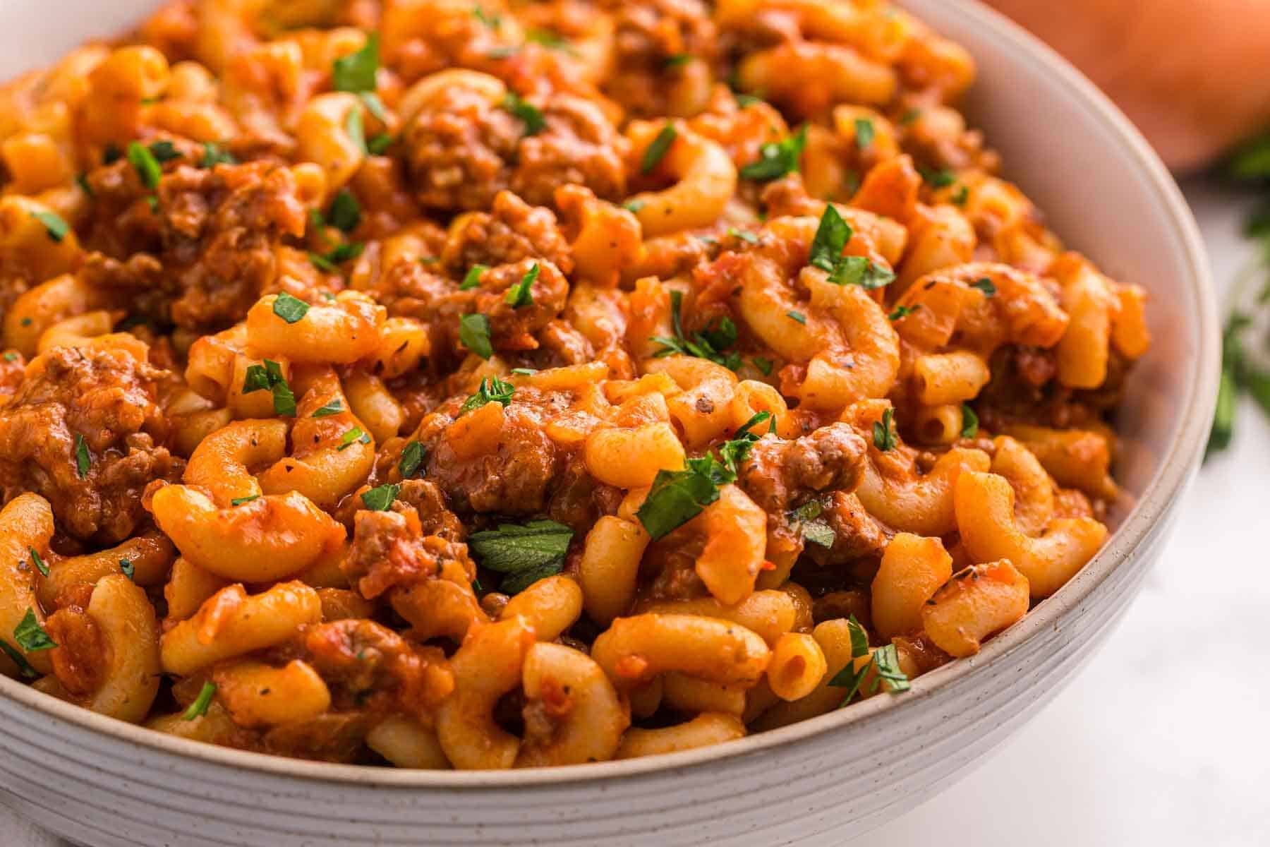 A close view of elbow macaroni and ground beef coated in thick tomato sauce and sprinkled with chopped parsley. The goulash is served in a wide ceramic bowl.