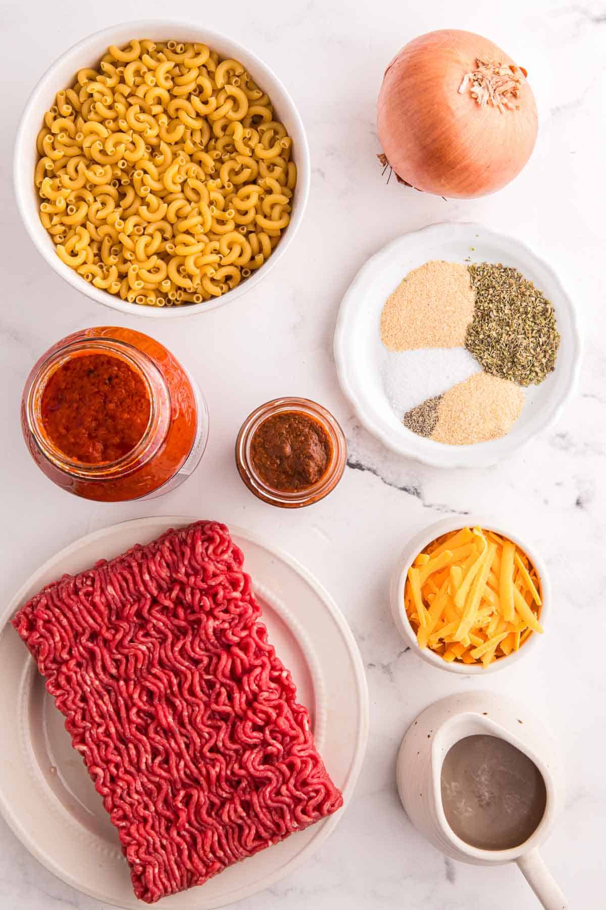 Ingredients for American goulash are arranged on a white counter. They include ground beef diced yellow onion marinara sauce petite diced tomatoes beef broth Italian seasoning garlic powder salt black pepper dry elbow macaroni and shredded sharp cheddar cheese.