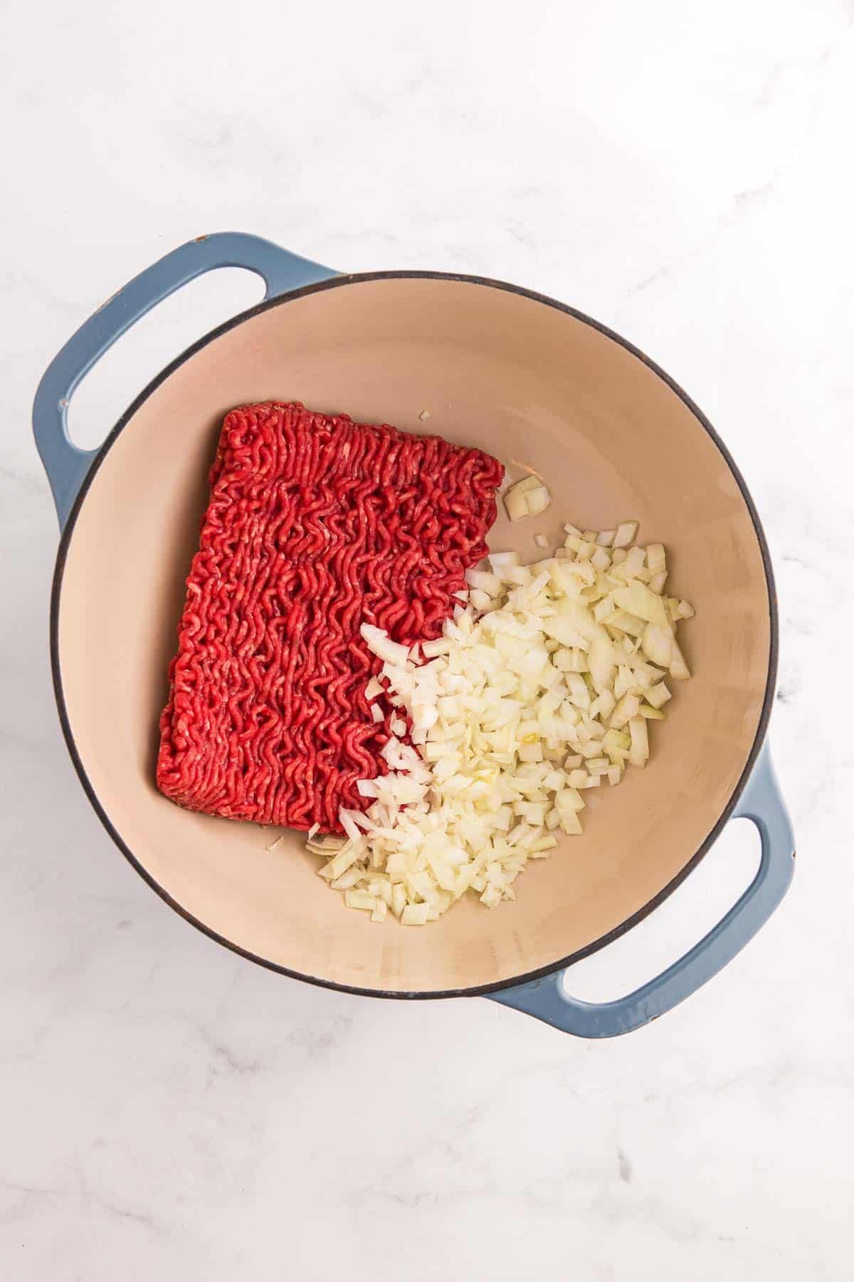 Raw ground beef and diced yellow onion sit together in a blue handled Dutch oven. This begins the first cooking step before the beef is browned and the onion softens.