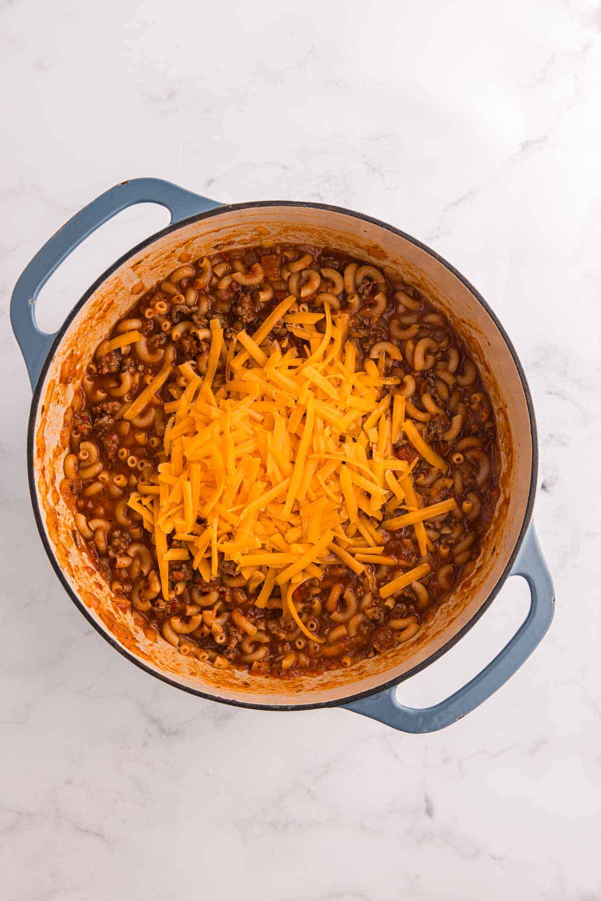 Shredded sharp cheddar cheese sits on top of the cooked macaroni beef and tomato mixture. Stirring it in will make the sauce creamy before the goulash rests.