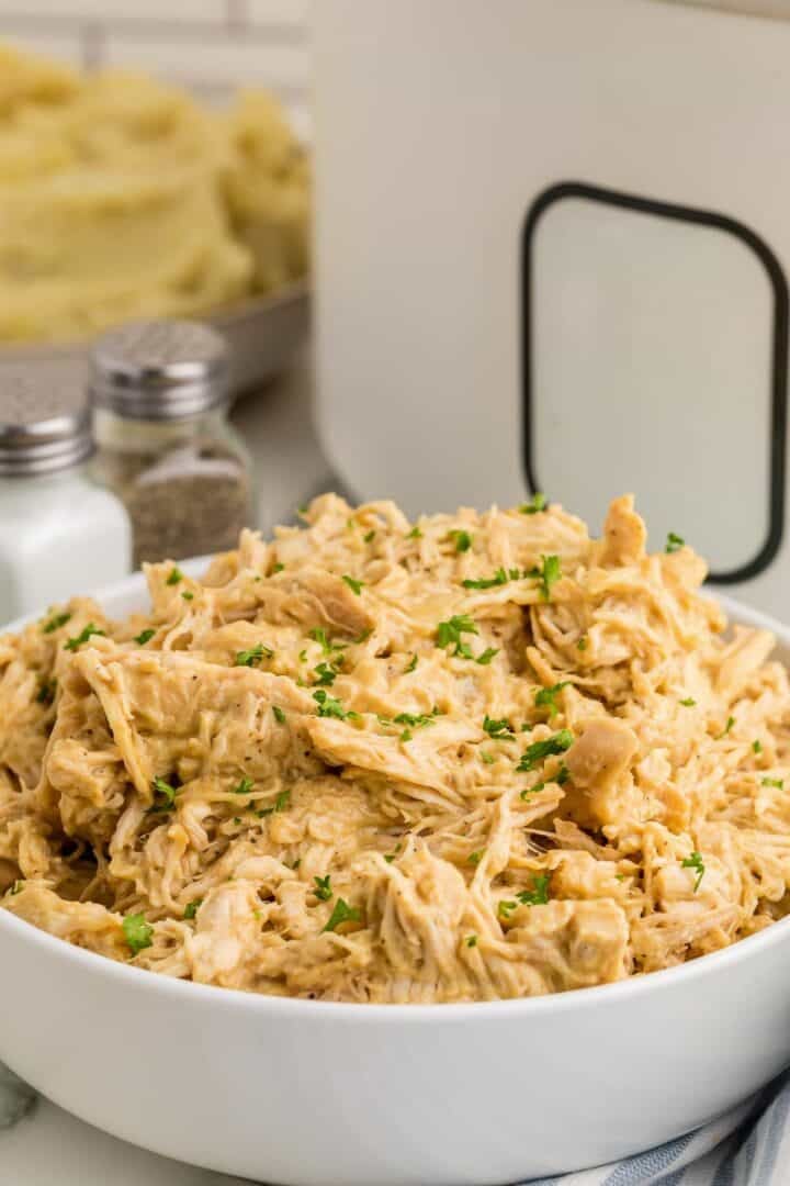 A serving bowl filled with Slow Cooker Honey Mustard Chicken and topped with chopped parsley. The shredded chicken is thoroughly coated in a thick golden sauce.