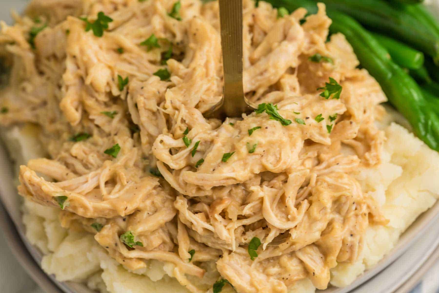 Close view of shredded honey mustard chicken piled over mashed potatoes with green beans alongside. A fork rests in the tender chicken.