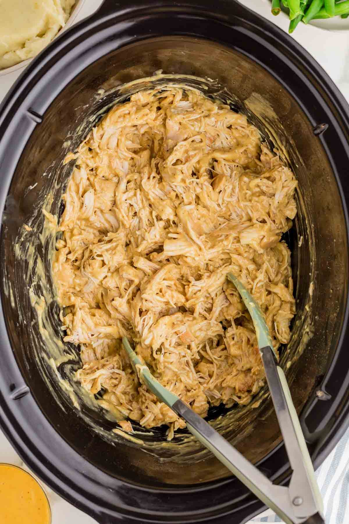 Tongs shred the cooked chicken directly in the slow cooker while mixing it into the thick honey mustard sauce. The chicken separates easily into tender strands.