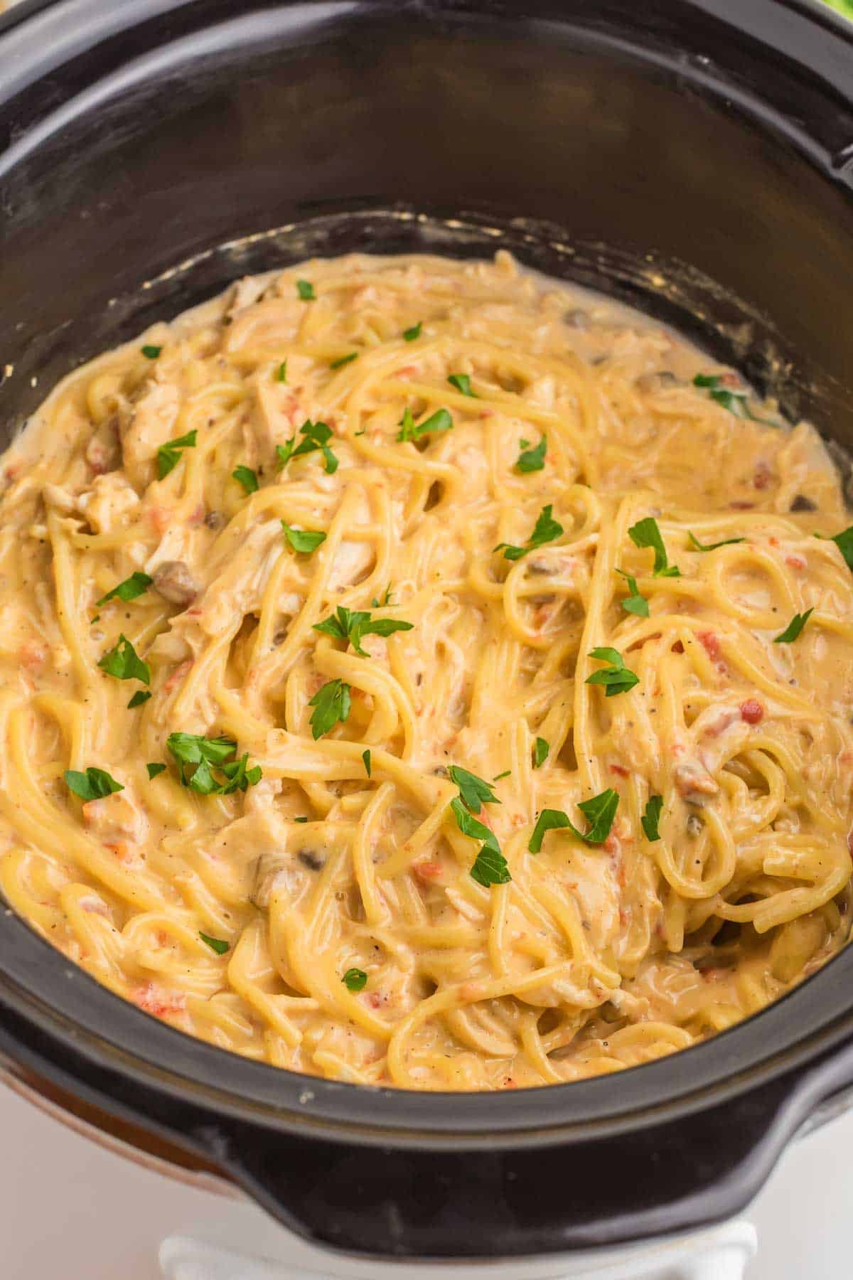 Rotel chicken spaghetti inside a slow cooker after the cooked pasta and shredded chicken have been stirred into the cheese sauce. Chopped parsley is scattered over the finished dish.