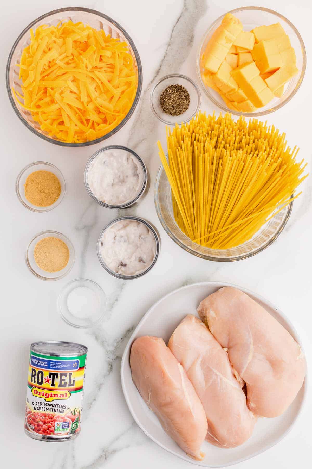 Ingredients for slow cooker Rotel chicken spaghetti arranged on a counter including chicken breasts, broken spaghetti, shredded cheddar cheese, cubed Velveeta, cream of mushroom soup, garlic powder, onion powder, salt, black pepper, and a can of ROTEL Original Diced Tomatoes and Green Chilies.
