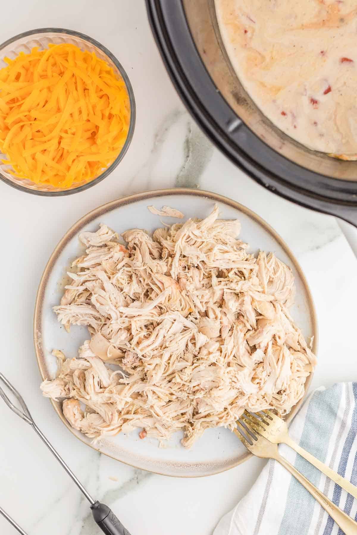 Cooked chicken shredded on a plate beside the slow cooker with shredded cheddar ready to be added to the sauce.