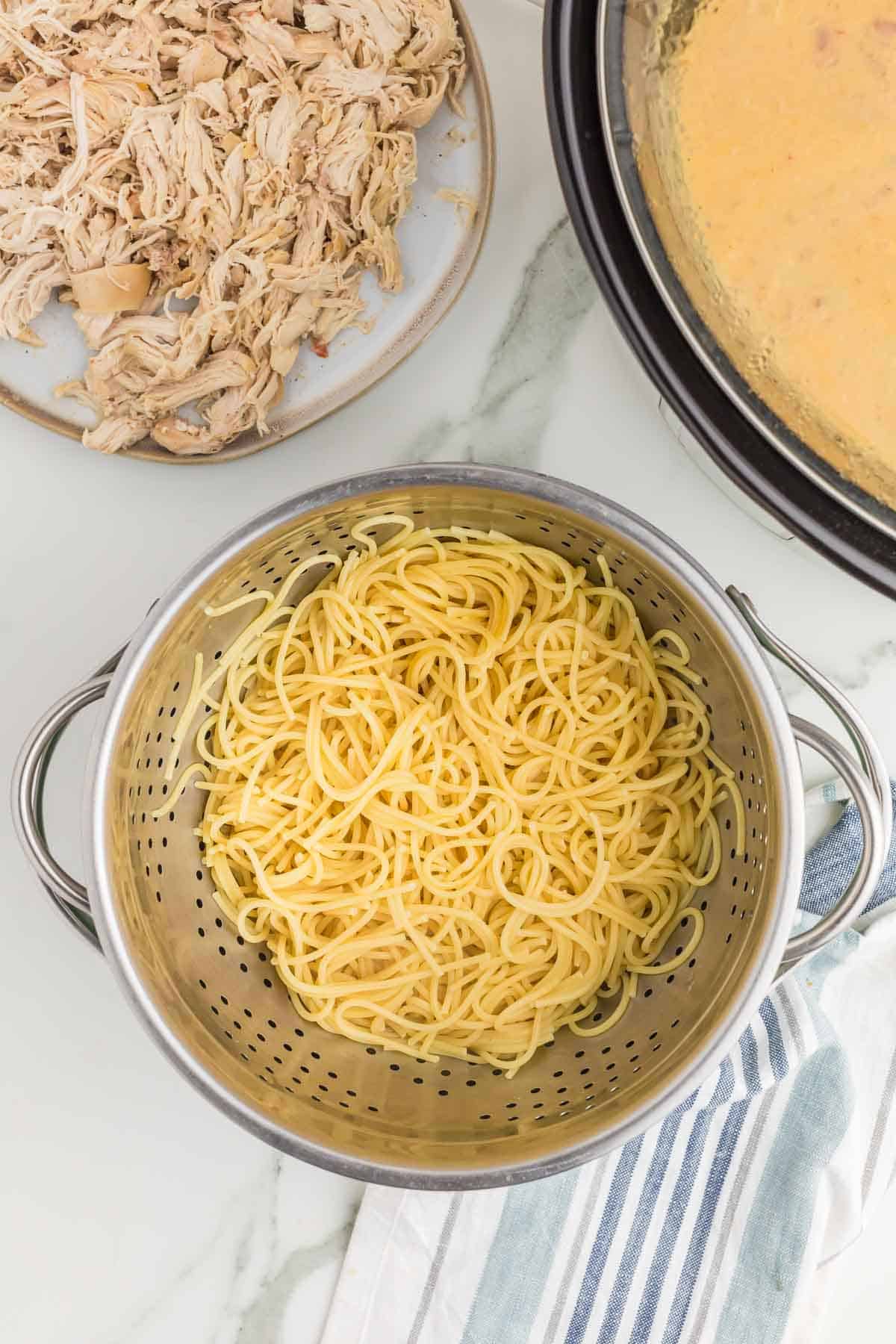 Cooked spaghetti draining in a colander beside shredded chicken and the prepared cheese sauce before everything is combined in the slow cooker.