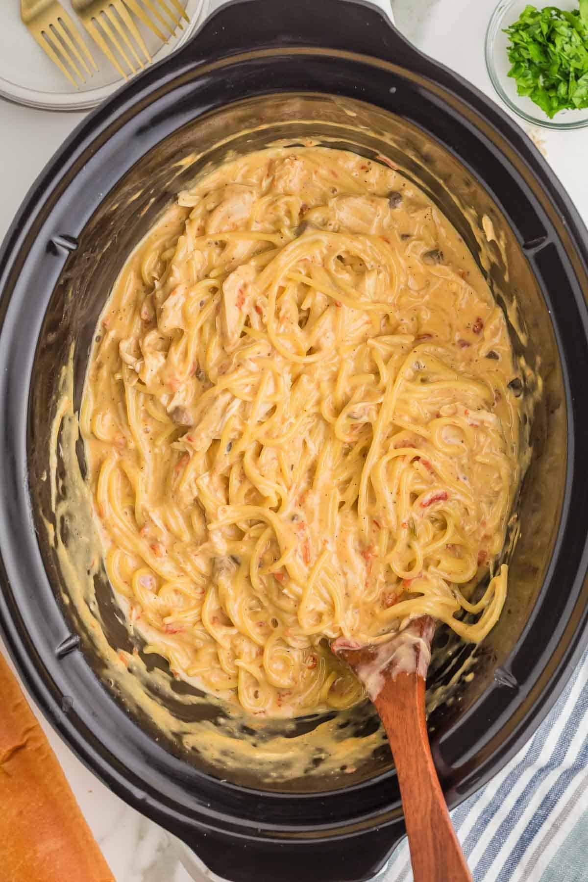 Cooked spaghetti and shredded chicken stirred into the Rotel cheese sauce inside the slow cooker with a wooden spoon resting in the mixture.