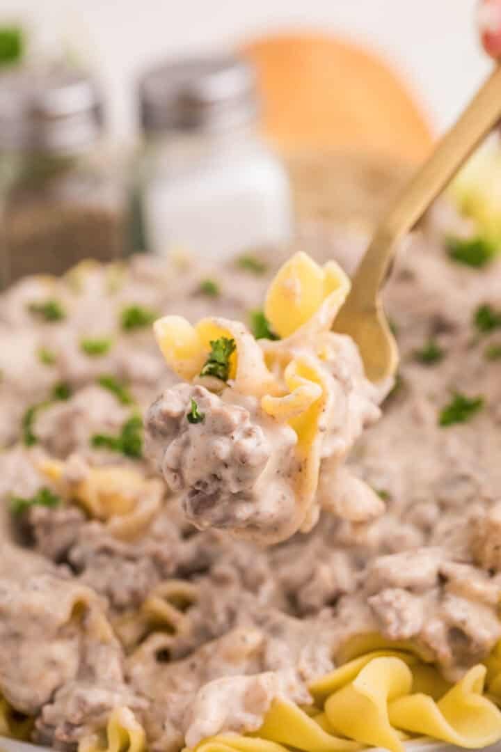 A fork lifts egg noodles coated in creamy ground beef stroganoff. Pieces of ground beef and parsley are visible in the sauce.