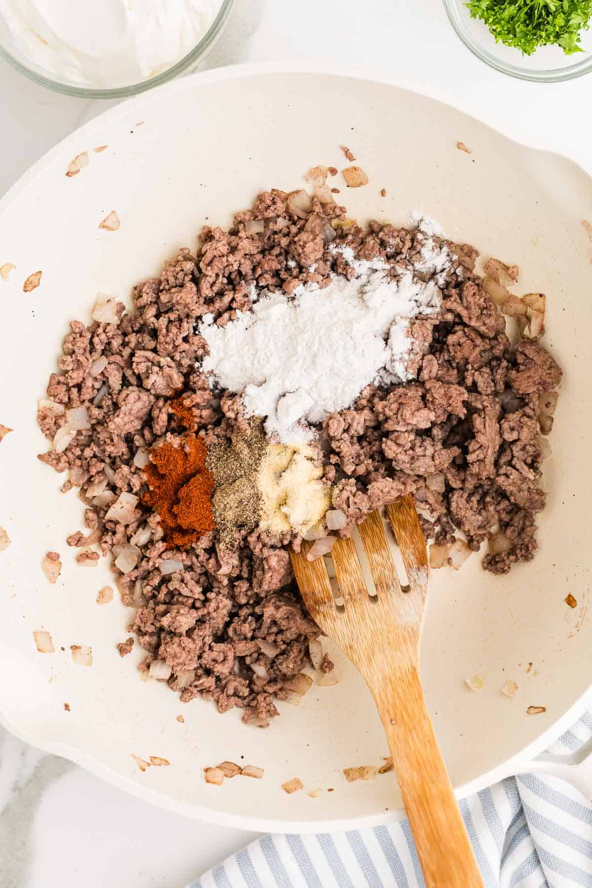 Flour salt garlic powder paprika and black pepper added over the cooked ground beef and onion. The seasonings are ready to be stirred into the beef.