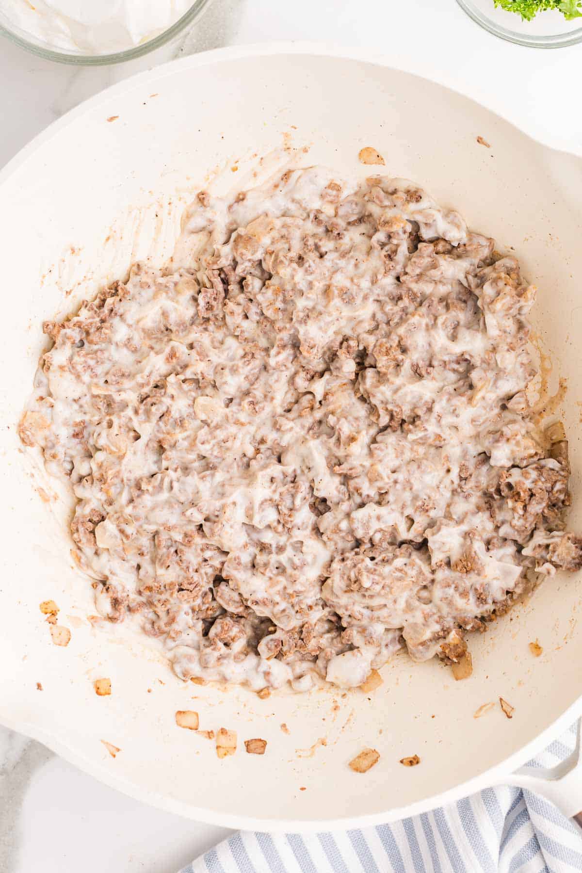 Ground beef stroganoff sauce cooking in the skillet after the creamy ingredients have been stirred in. The sauce is thick and coats the crumbled beef and onion.