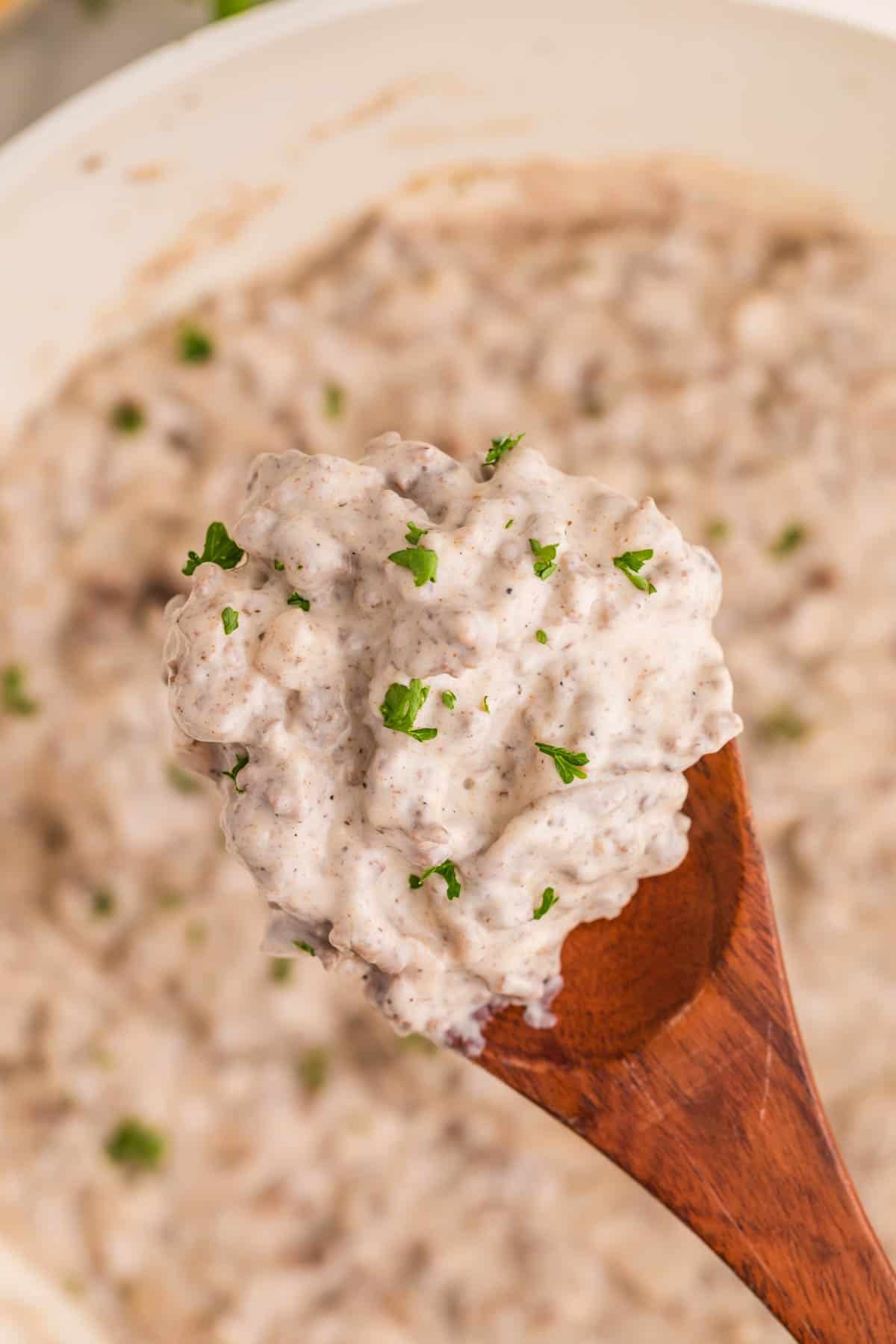 A wooden spoon holds a scoop of finished ground beef stroganoff above the skillet. The creamy sauce is thick with crumbled beef and topped with parsley.