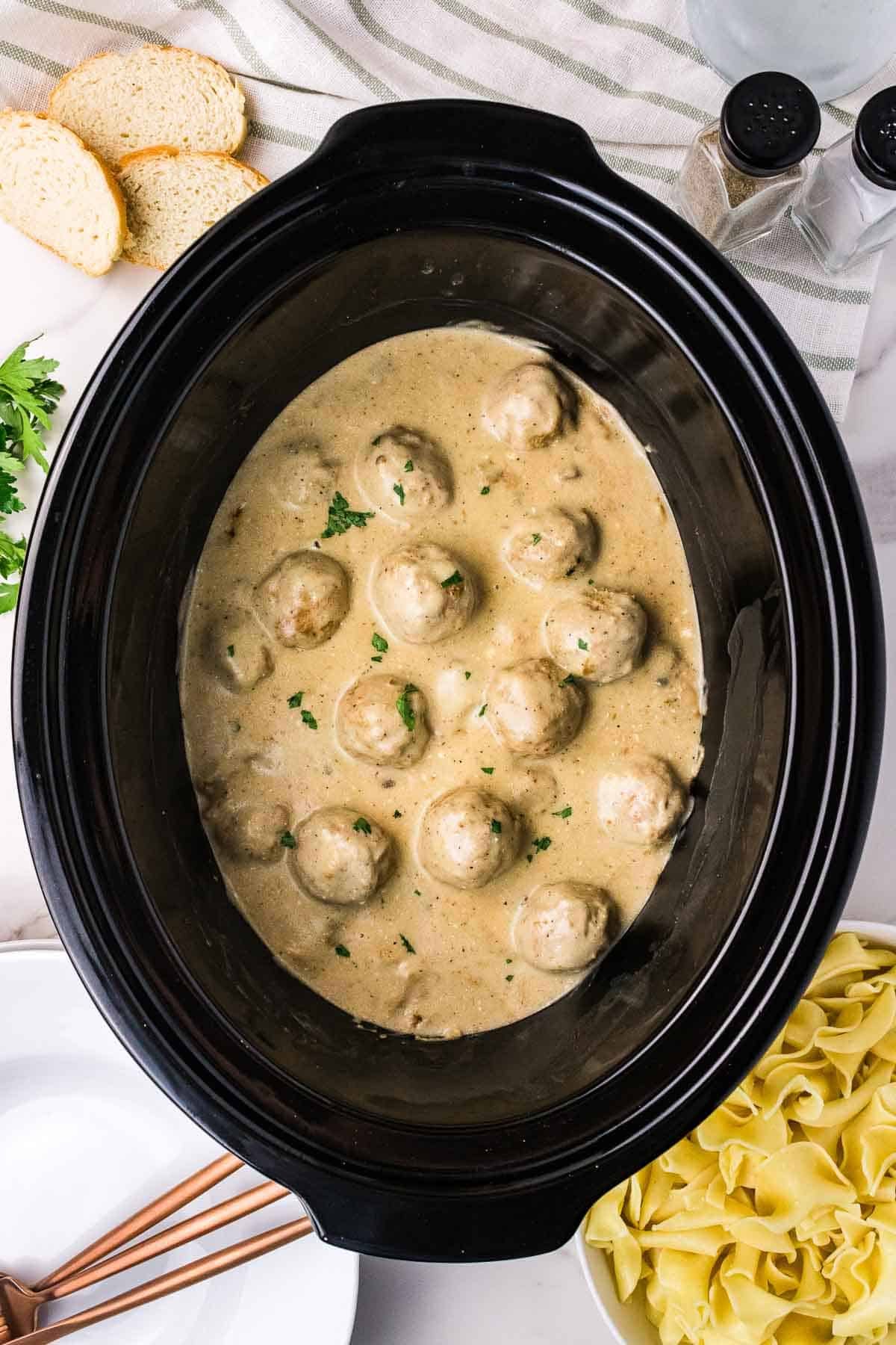 Finished Crockpot Swedish meatballs in creamy mushroom sauce inside the slow cooker. Chopped parsley is sprinkled over the meatballs before they are served with egg noodles.
