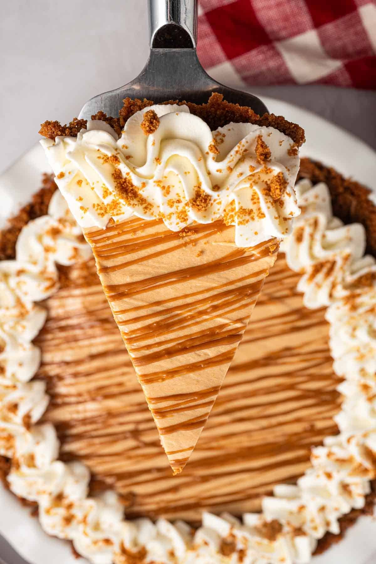 Slice of no bake Biscoff cookie butter pie being lifted from the whole pie with a pie server. Whipped cream and crushed Biscoff cookies line the crust while cookie butter is drizzled across the filling.