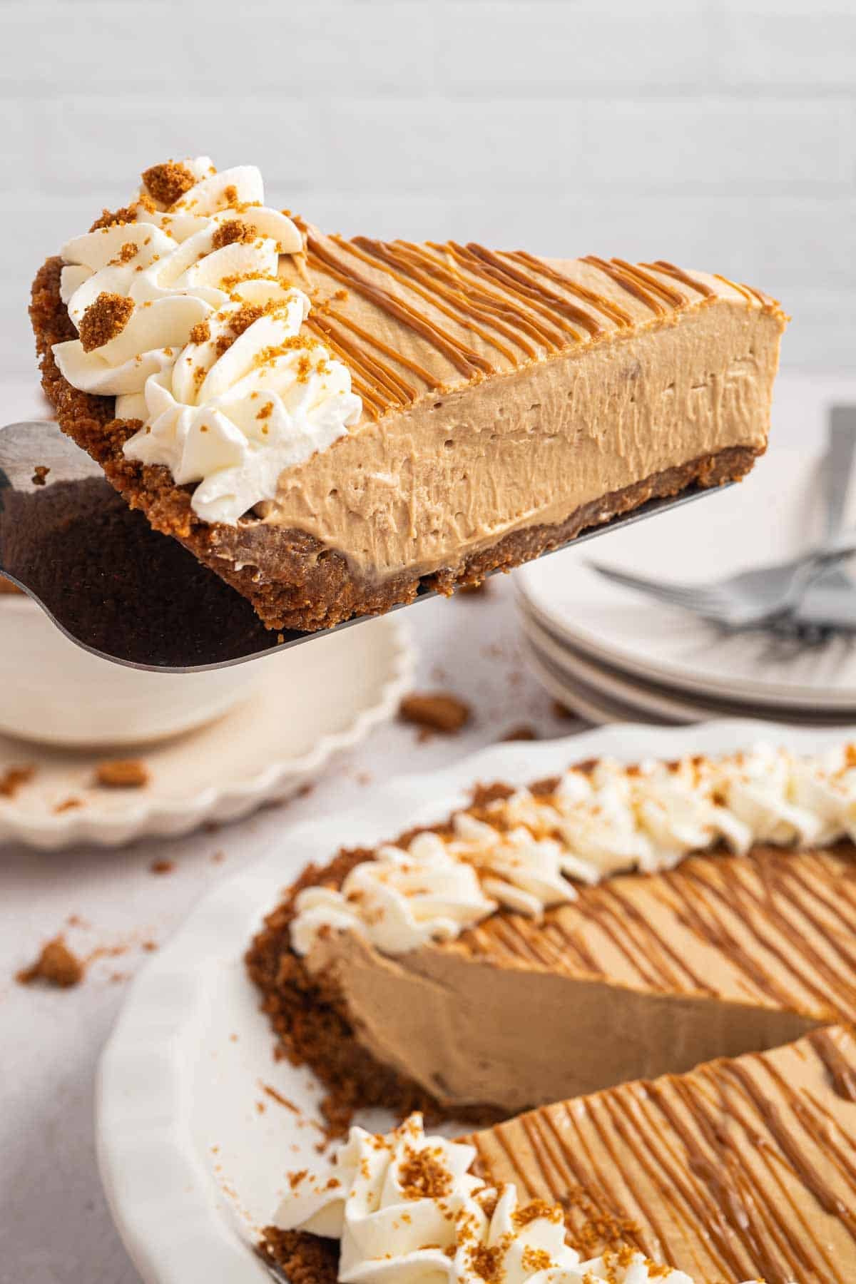 Side view of a slice of Biscoff cookie butter pie on a pie server. The thick cookie butter filling sits over a dark cookie crumb crust and is topped with whipped cream and cookie crumbs.