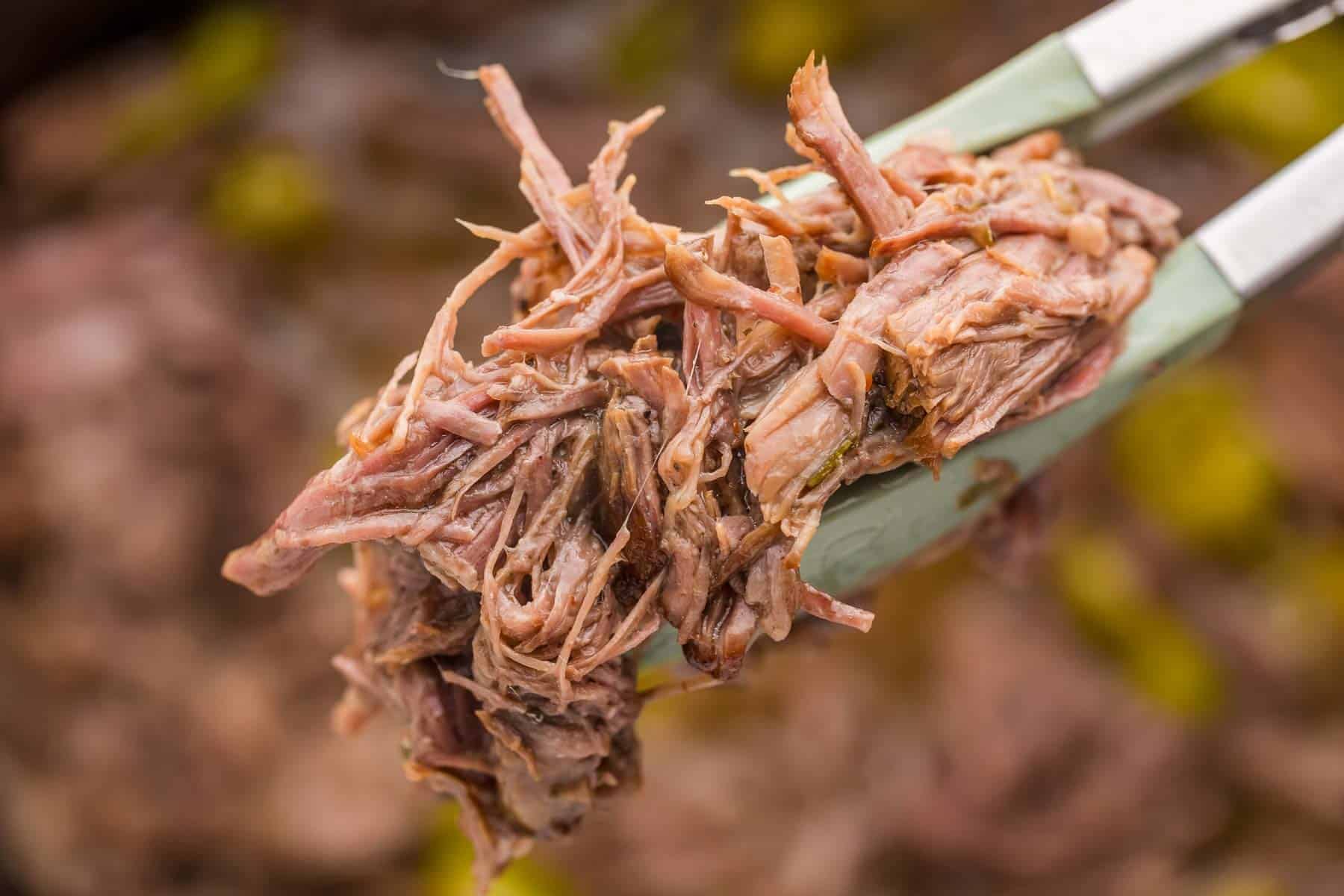 Silicone tipped tongs lift a close up portion of tender shredded pot roast from the slow cooker.