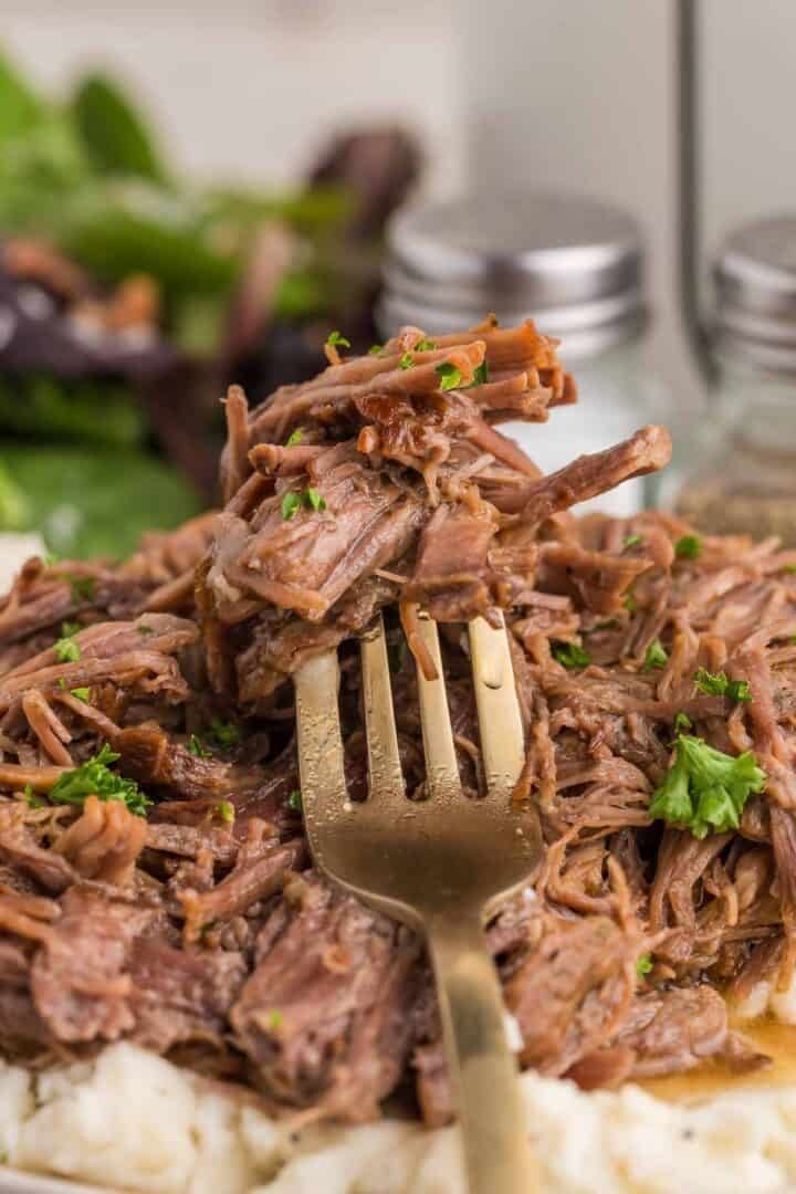 A fork lifts a bite of shredded pot roast above the plated beef and mashed potatoes. The close view shows the soft pulled texture of the slow cooked meat.