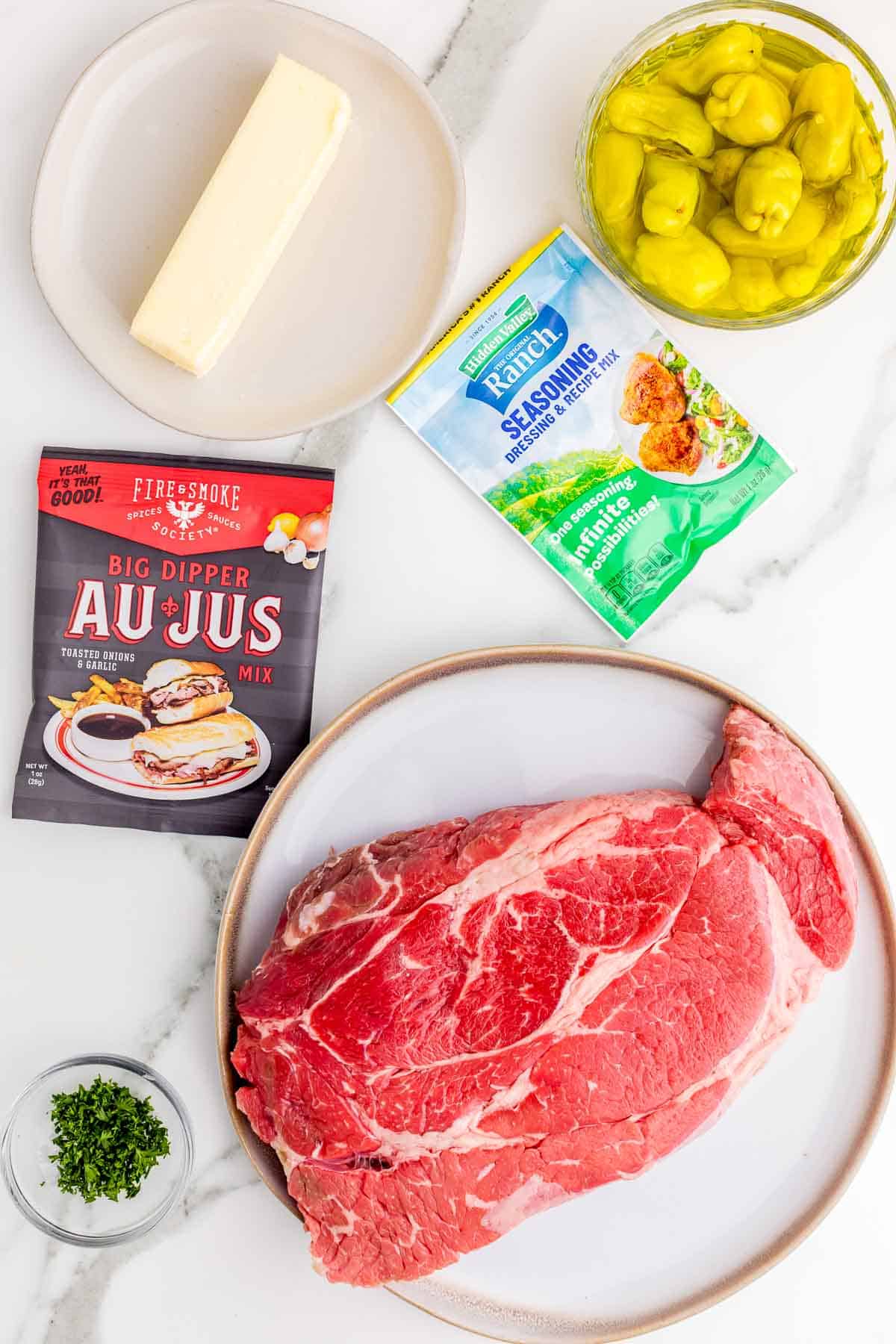Ingredients for Mississippi pot roast arranged on a white marble surface with a raw beef chuck roast, butter, whole pepperoncini peppers, chopped parsley, a ranch mix packet, and an au jus packet.