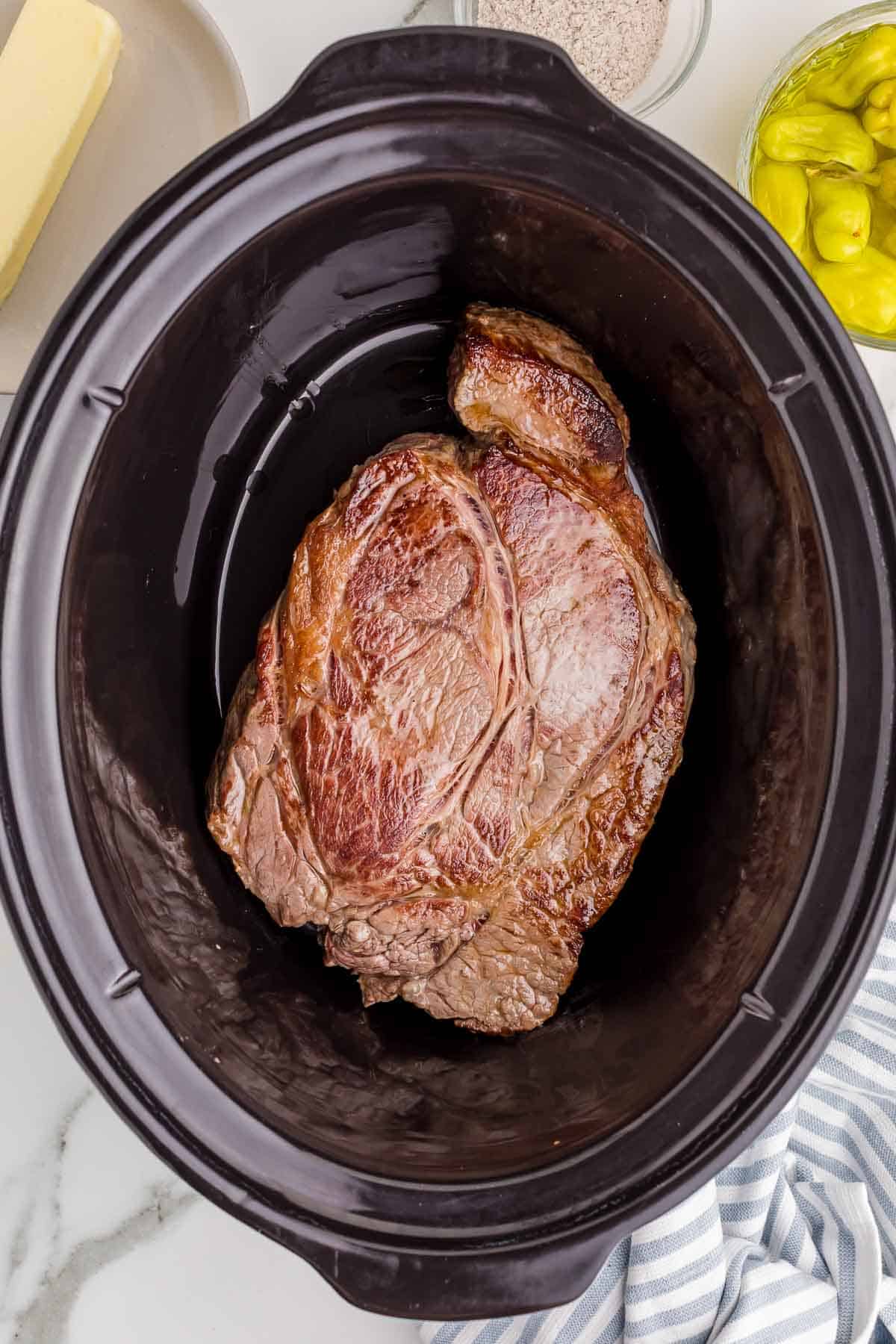 A browned beef chuck roast rests in the bottom of a black slow cooker after being seared.
