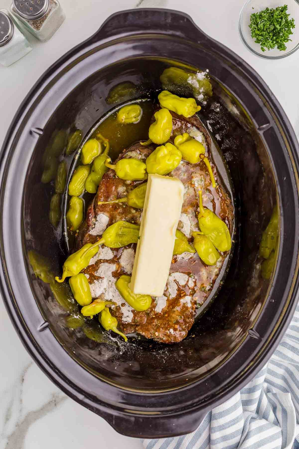 The seared chuck roast is topped with ranch seasoning and au jus mix in the slow cooker. Whole pepperoncini peppers and their liquid surround the beef with a stick of butter placed across the center before cooking.