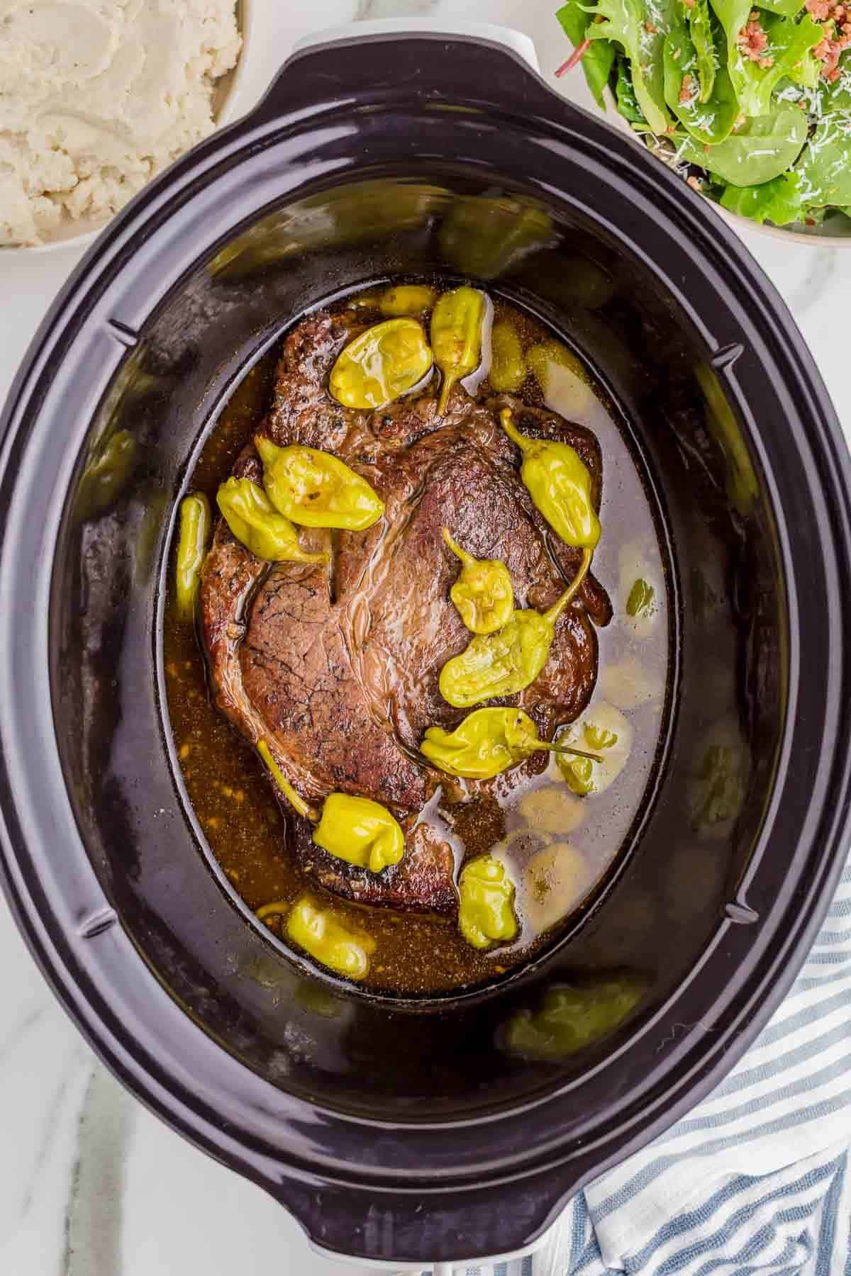 Cooked Mississippi pot roast rests in its cooking juices with softened pepperoncini peppers scattered across the top.