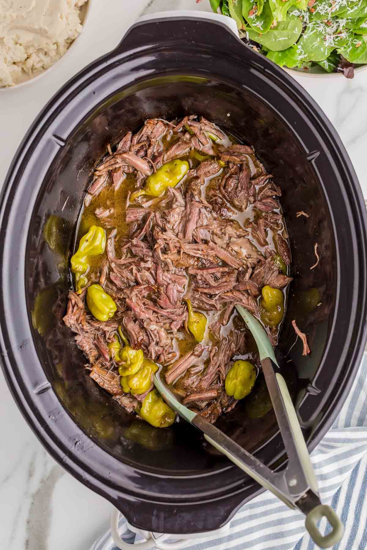 The finished pot roast has been shredded directly in the slow cooker and mixed with the cooking juices and pepperoncini peppers. Two silicone tipped tongs rest in the tender beef ready for serving.