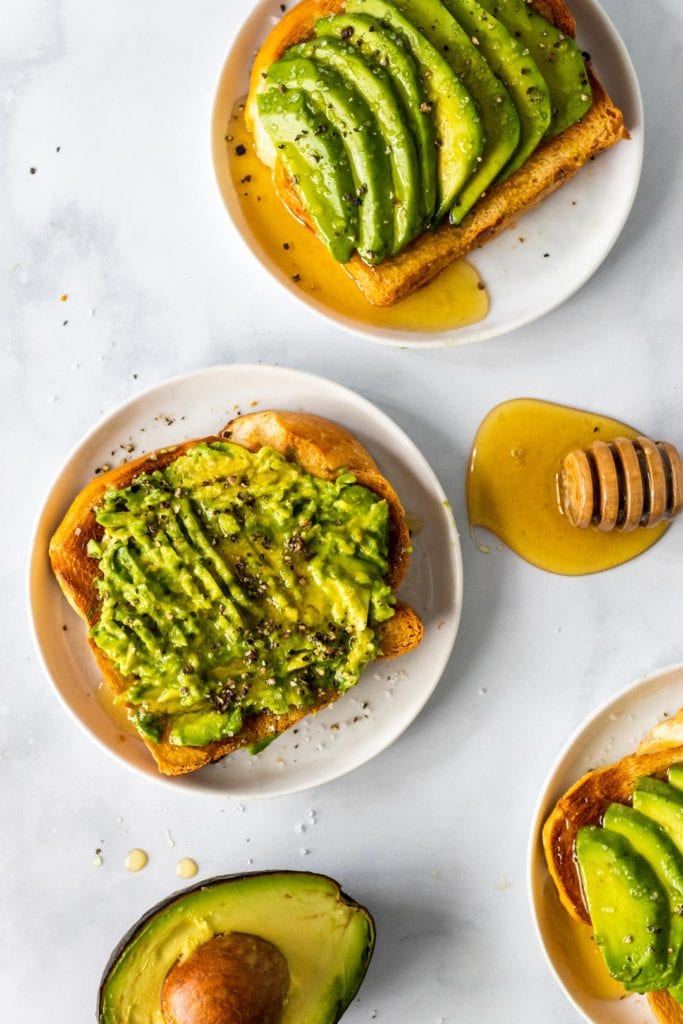 A slice of avocado toast topped with honey, sea salt and course grind black pepper; a wooden honey dipper is off to the size in a pool of honey