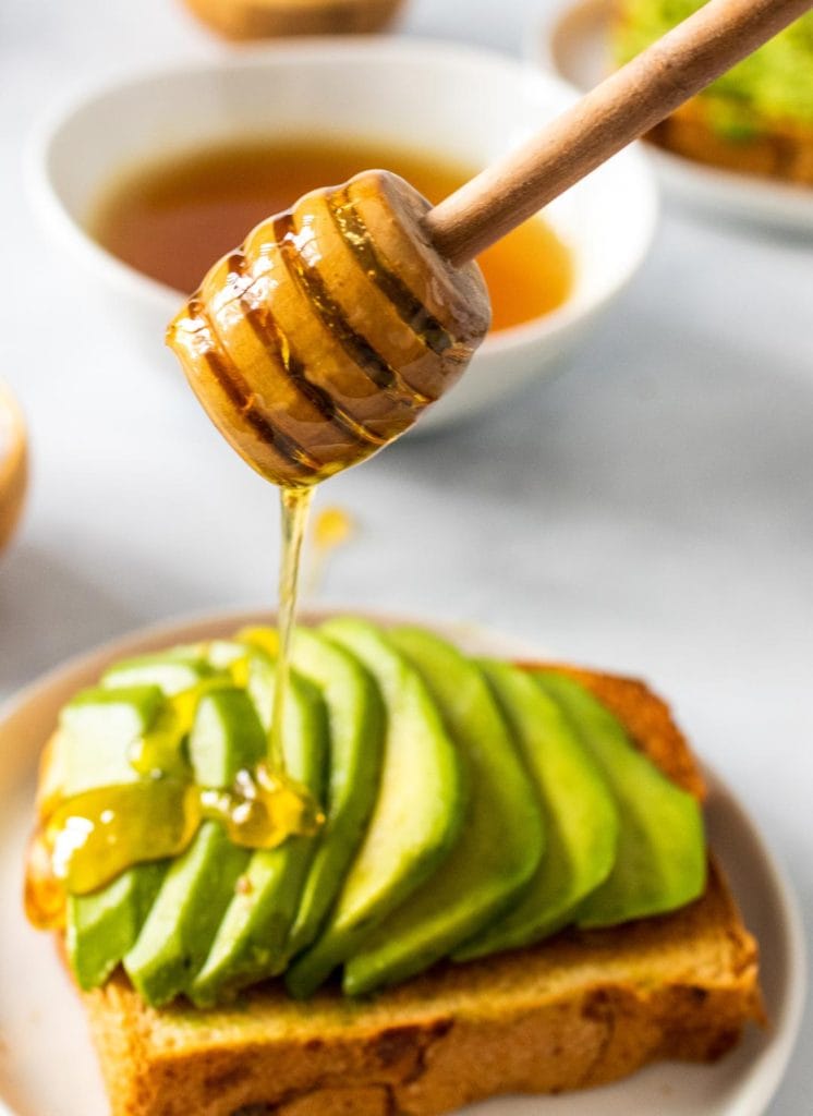 Honey being drizzled from a wooden honey dipper over slices of avocado atop a piece of toast